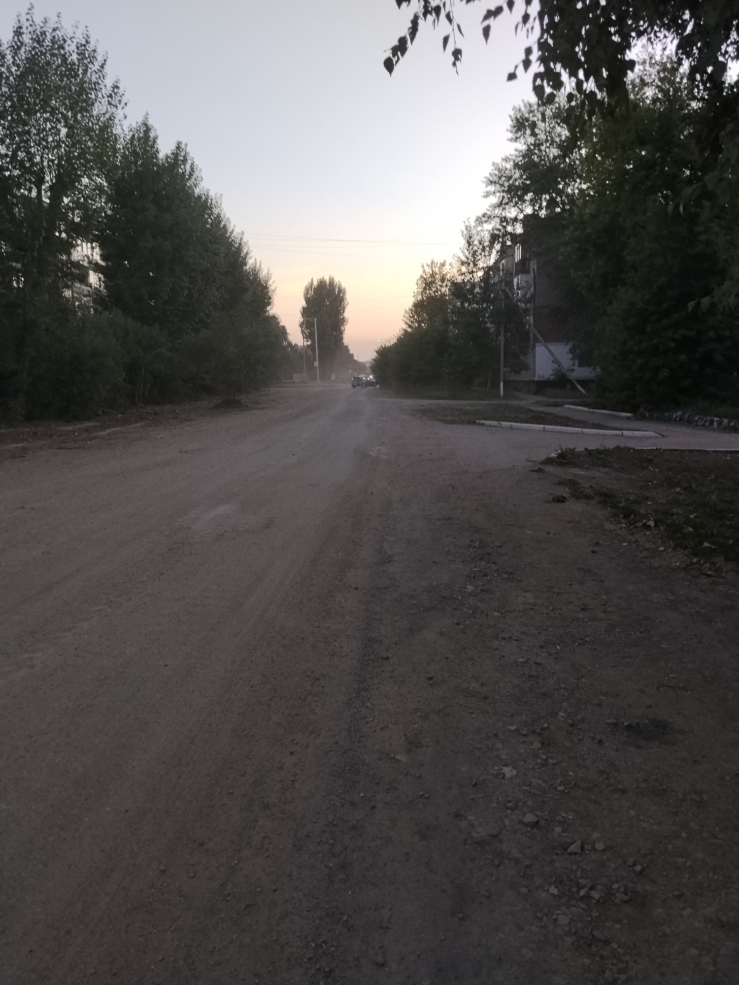The image shows a dirt road in a residential area of Kokshetau, Kazakhstan. The road is unpaved and appears to be in disrepair, with visible potholes and loose gravel. On either side of the road are trees and buildings, including what appears to be a multi-story apartment building on the right. The sky is overcast, and the lighting suggests it is either early morning or late evening. There are no people visible in the image, but there is a parked vehicle in the distance on the road. The overall scene depicts a quiet, somewhat neglected urban environment.