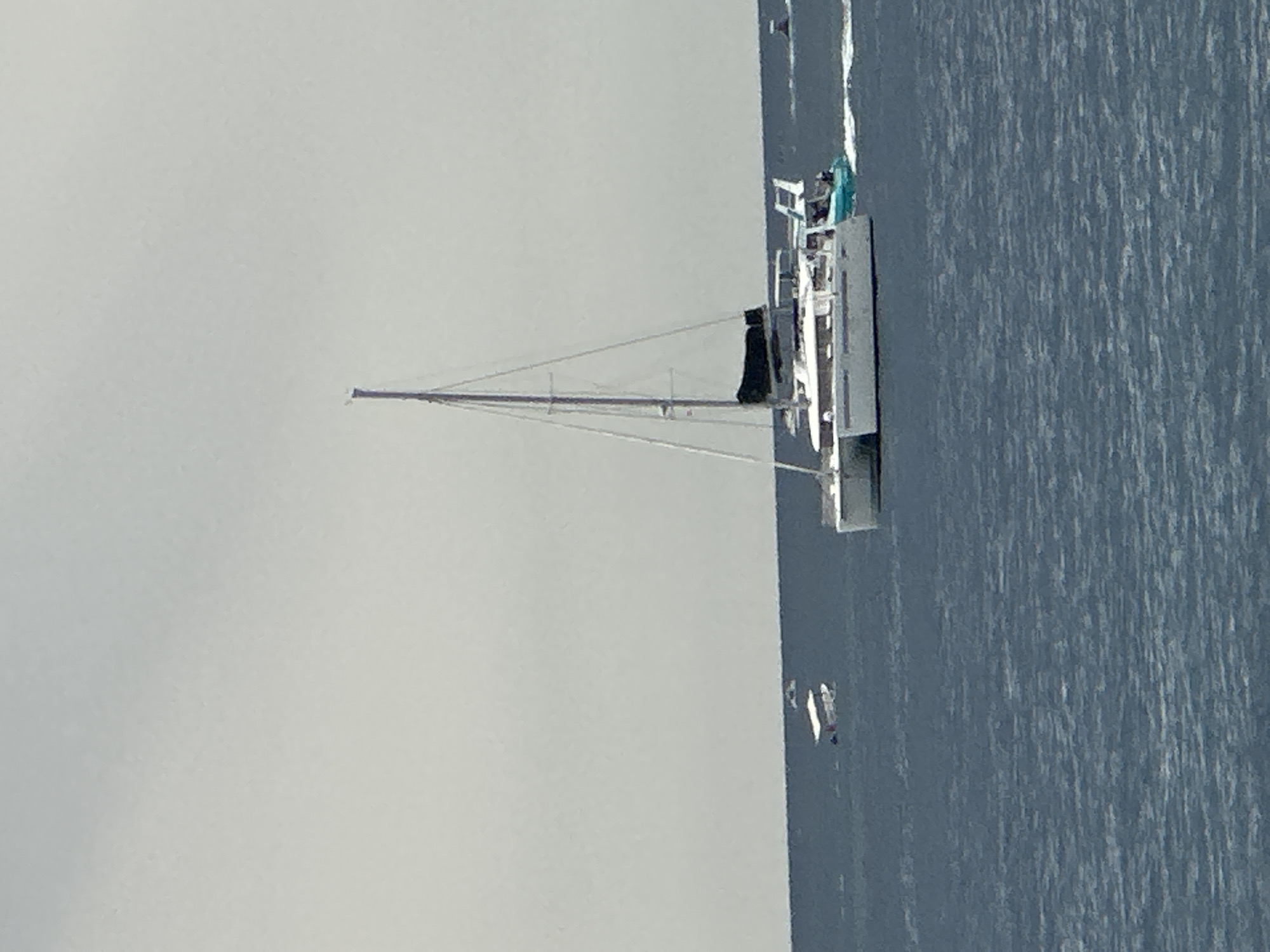A wide, horizontal, medium-distance shot captures a cloudy, gray sky over a vast, choppy, dark blue ocean. In the foreground, the ocean's surface is textured with many small waves. On the right side of the image, a large white catamaran with two hulls and a tall mast is sailing. It has a black sail furled at the top of the mast and white cabin structures on its decks. A smaller, teal-colored boat is positioned closely behind the catamaran, leaving a white wake. To the left of the catamaran, further in the distance, are two smaller, white boats. The boat on the left appears to be a dinghy with occupants visible. In the far distance on the right, another small boat is visible, also leaving a wake. The horizon line is barely discernible where the water meets the overcast sky. The scene is set at sea, likely near the coast, as indicated by the presence of multiple vessels. The image does not provide specific details to pinpoint the exact location in Budva, Montenegro, but it depicts a typical maritime activity in coastal waters.