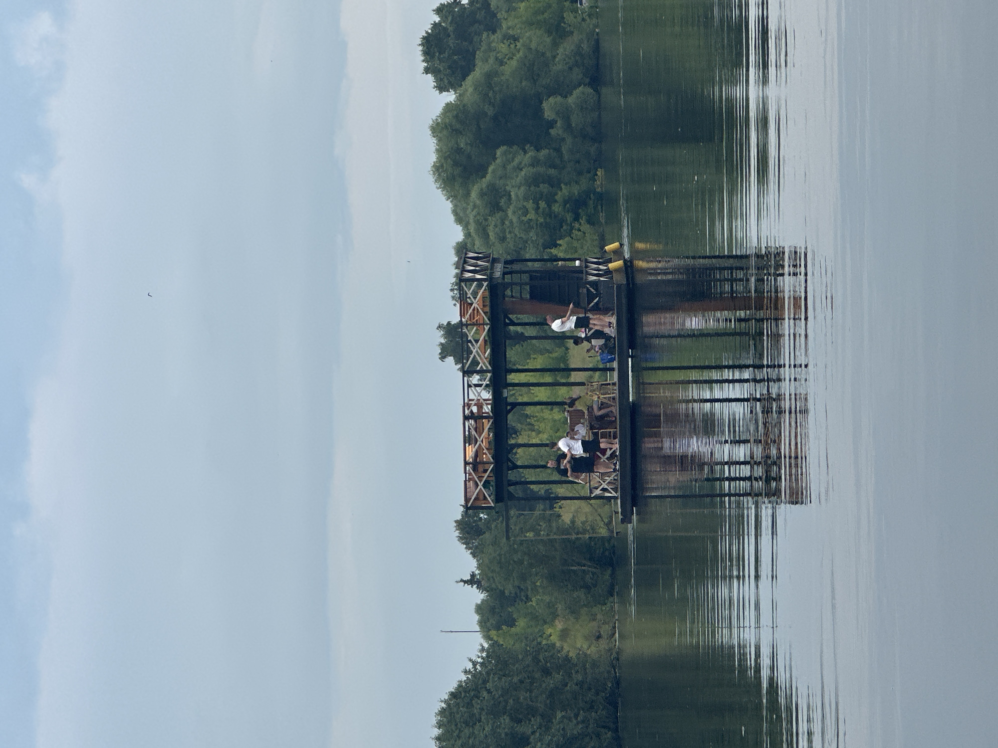The image depicts a floating structure with a multi-level wooden deck on a body of water, possibly a lake or river. The structure is occupied by several individuals, some of whom appear to be socializing or engaged in activities. The background reveals a densely forested shoreline under an overcast sky. A lone bird is visible in the sky. The scene reflects on the calm water surface, indicating minimal wind. The location is identified as the city of Gorki Leninskiye, Russia.