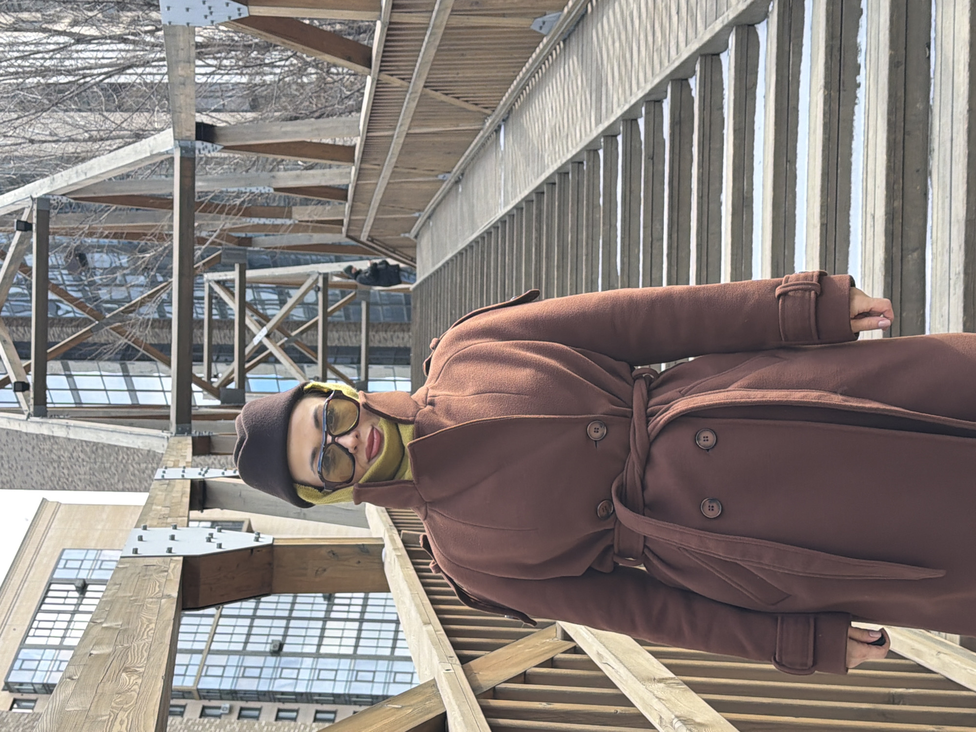 A woman in a brown coat, hat, and yellow scarf is posing on a pedestrian bridge. The bridge is made of light-colored wood and concrete, with railings on one side. In the background, there are buildings with glass facades and bare tree branches. The location is likely a park or public space in Krasnoyarsk, Russia. The image is rotated 90 degrees counter-clockwise.
