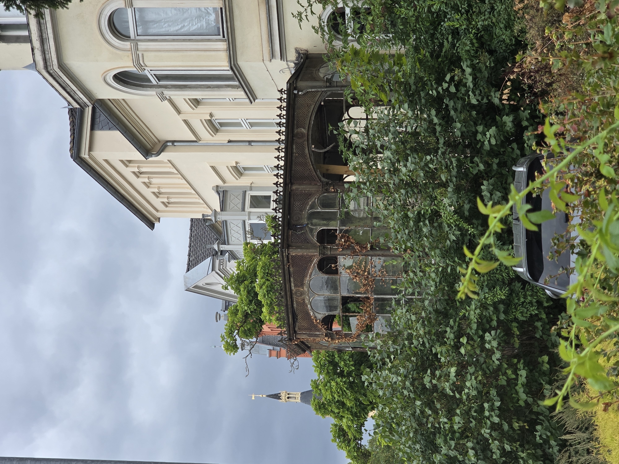 The image shows a view of old buildings in Schwerin, Germany, on a cloudy day. In the foreground, lush green bushes obscure the lower part of the scene, with a dark-colored car partially visible beneath them. Behind the bushes, an ornate, derelict conservatory with large glass windows and a rusted metal frame is prominent. The conservatory is overgrown with vines and dry leaves.
Behind the conservatory, several buildings are visible. To the left, a tall, dark tower with a gold spire and cross rises above the greenery, suggesting it is part of a church or cathedral. Next to it are the red and cream-colored facades of older buildings. To the right, a larger, cream-colored building with arched windows and decorative architectural details is seen. The sky is overcast with heavy grey clouds.
