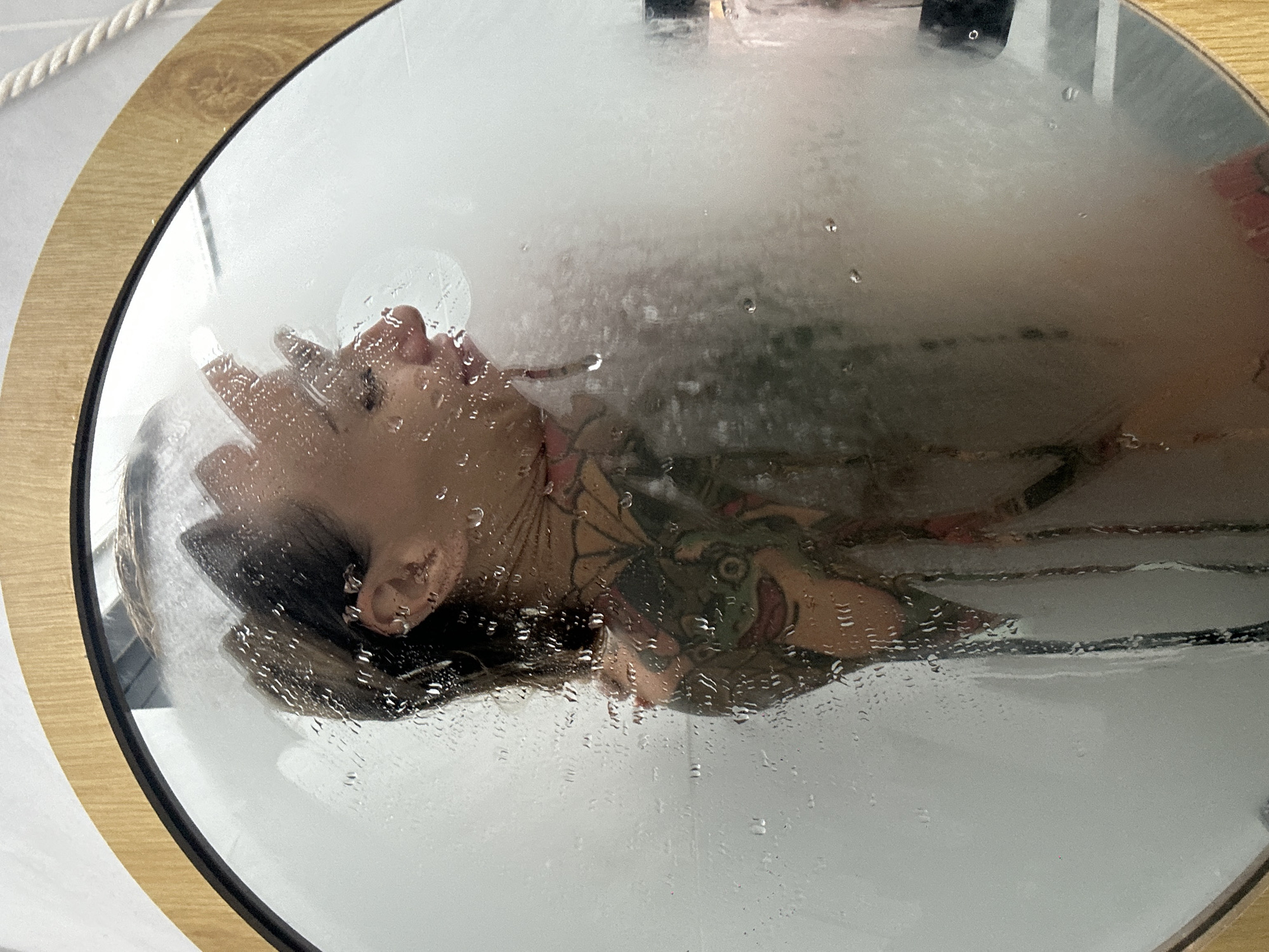 A woman with tattoos is reflected in a steamy, water-speckled mirror. She is wearing a bikini. The scene appears to be a bathroom. The image does not contain any information about the city of Mũi Né, Vietnam.