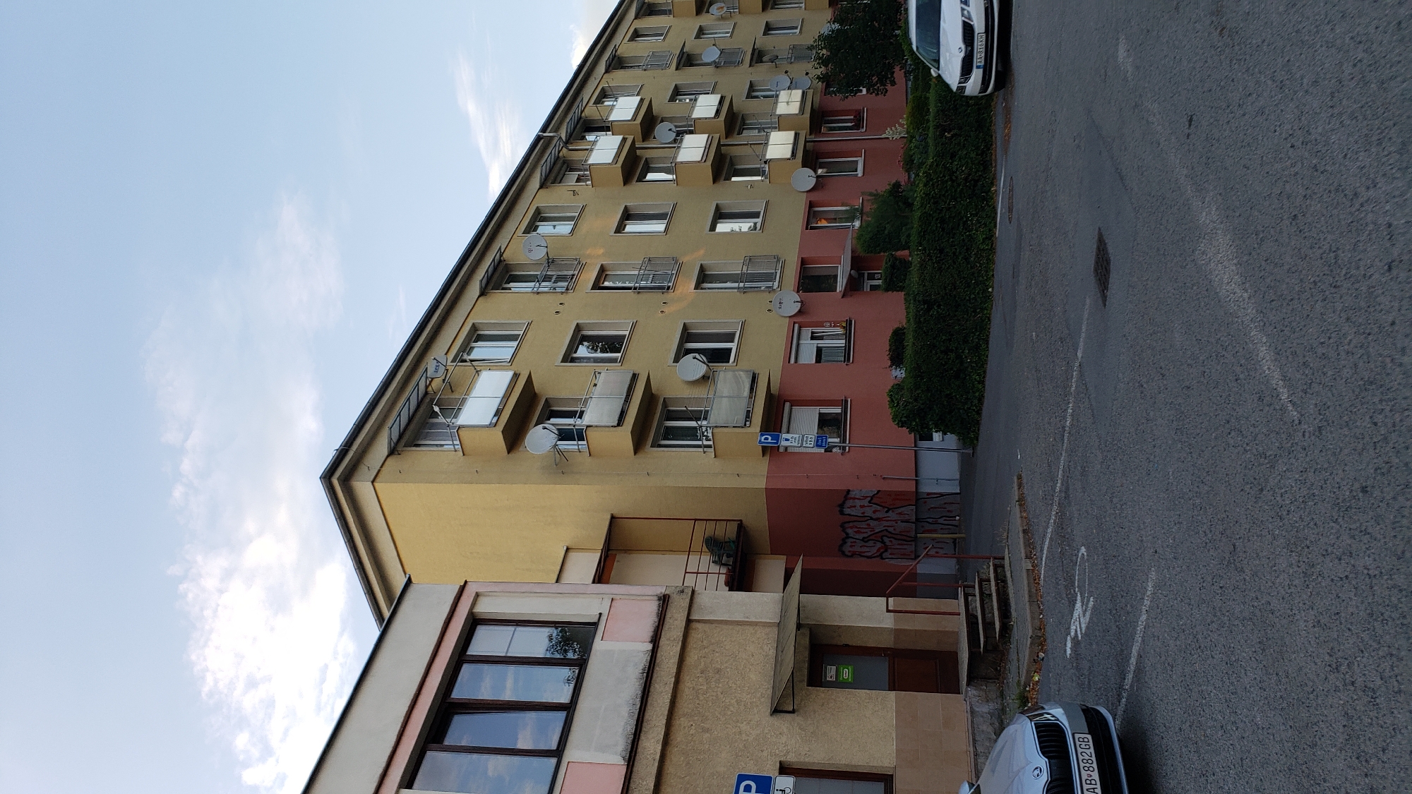 This image captures an exterior view of a yellow and pink multi-story apartment building in Prešov, Slovakia. The building features numerous balconies with white railings and satellite dishes mounted on the exterior walls. A section of the lower part of the building is painted pink and adorned with graffiti. To the left, a smaller building with beige and pink accents is visible, with a dark-colored door and a small awning. In front of the buildings, a paved area serves as a parking lot with designated parking spaces marked by white lines. A silver car with the license plate AB 882GB is parked to the left, and a white car with the license plate AA 816KH is parked on the right. A disabled parking symbol is painted on the asphalt in front of the silver car. Green bushes line the front of the apartment building. The sky above is a light blue with scattered clouds, suggesting it is daytime. The scene appears to be a residential area in the city.
