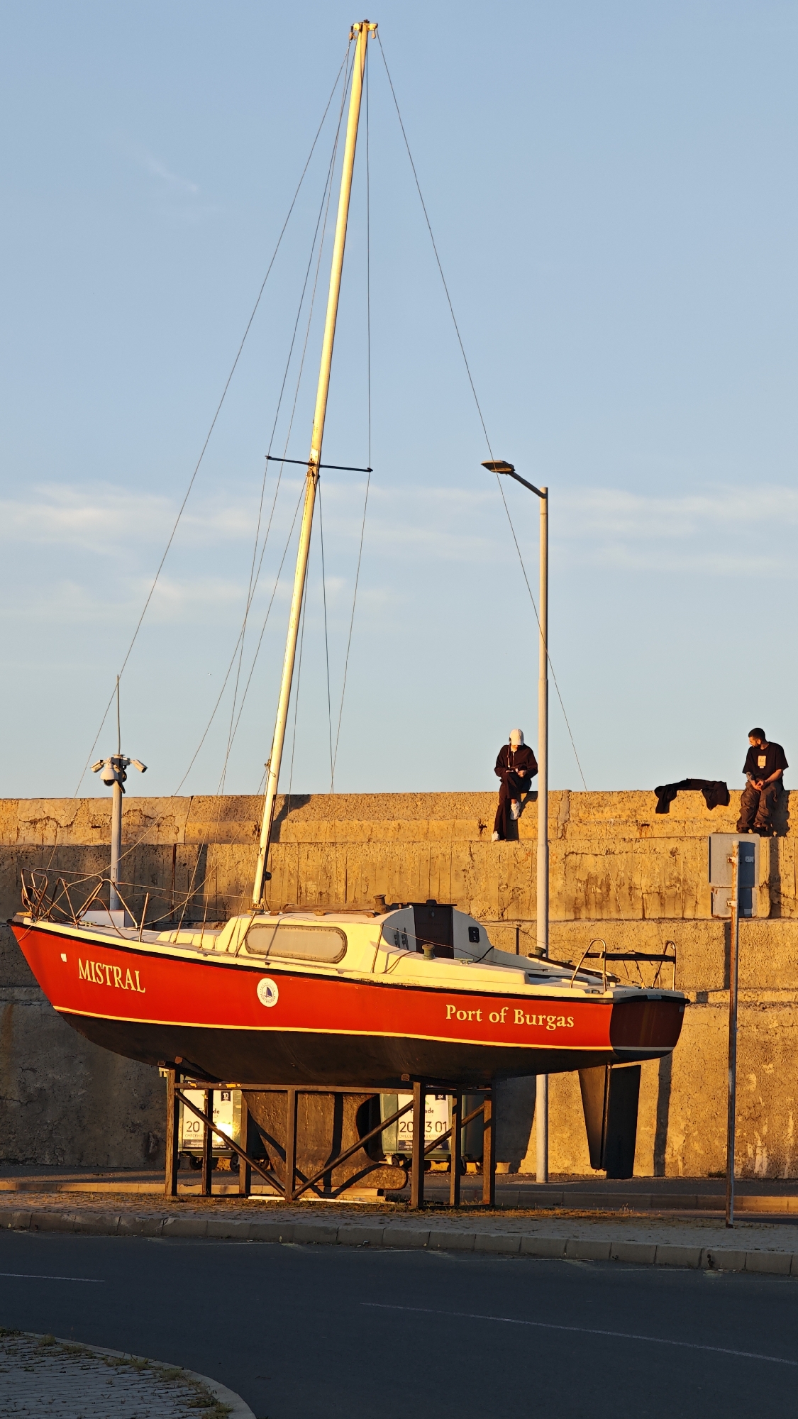 The image captures a sailboat named "Mistral Port of Burgas" resting on a black metal stand near a street. The boat has a red hull with a yellow top and a tall, slender mast extending upwards. Two men are sitting on a concrete wall behind the boat, under the light of a tall street lamp. A piece of clothing or material lies beside one of them. The wall appears to be part of a harbor or waterfront structure. The setting sun casts a warm light on the wall, boat, and surrounding objects, suggesting either sunrise or sunset. The street in front of the boat is asphalt. The scene appears to be in Burgas, Bulgaria, as indicated by the name on the boat.