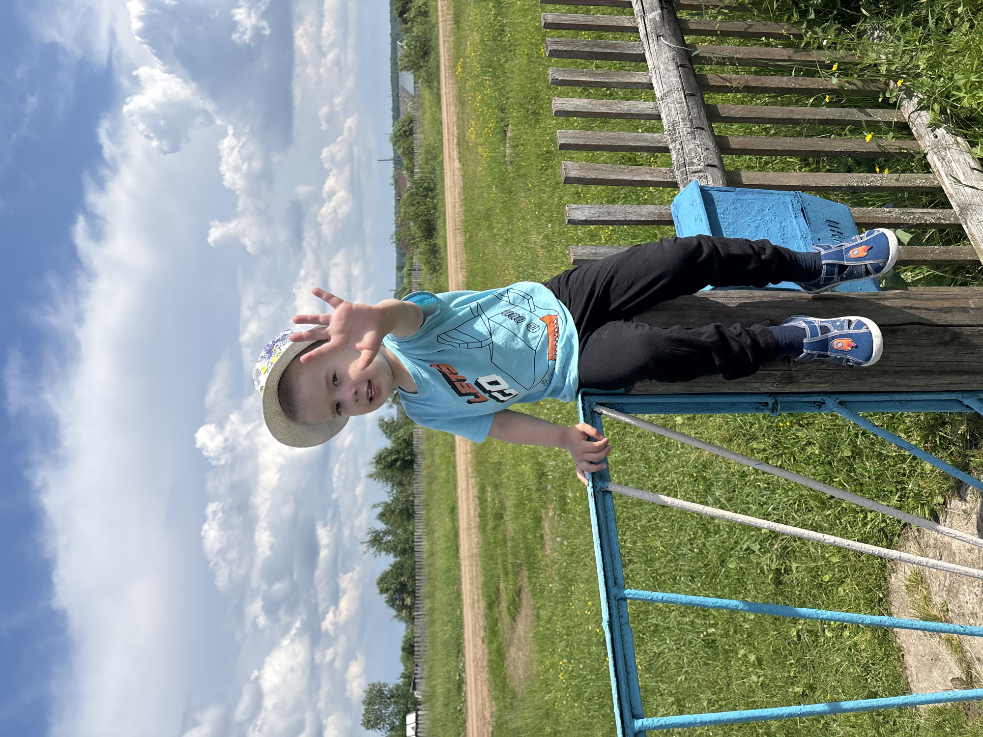 A young Caucasian child wearing a blue t-shirt with a graphic print and black pants sits on a rustic wooden fence. The child has on a straw hat and blue sandals with rocket designs. The child's right hand is raised, palm facing the camera. The background features a grassy field with a dirt road, trees, and a few houses in the distance under a cloudy blue sky. The scene appears to be in a rural area, possibly on the outskirts of the city of Kurgan, Russia. A blue mailbox is attached to the fence near the child.
