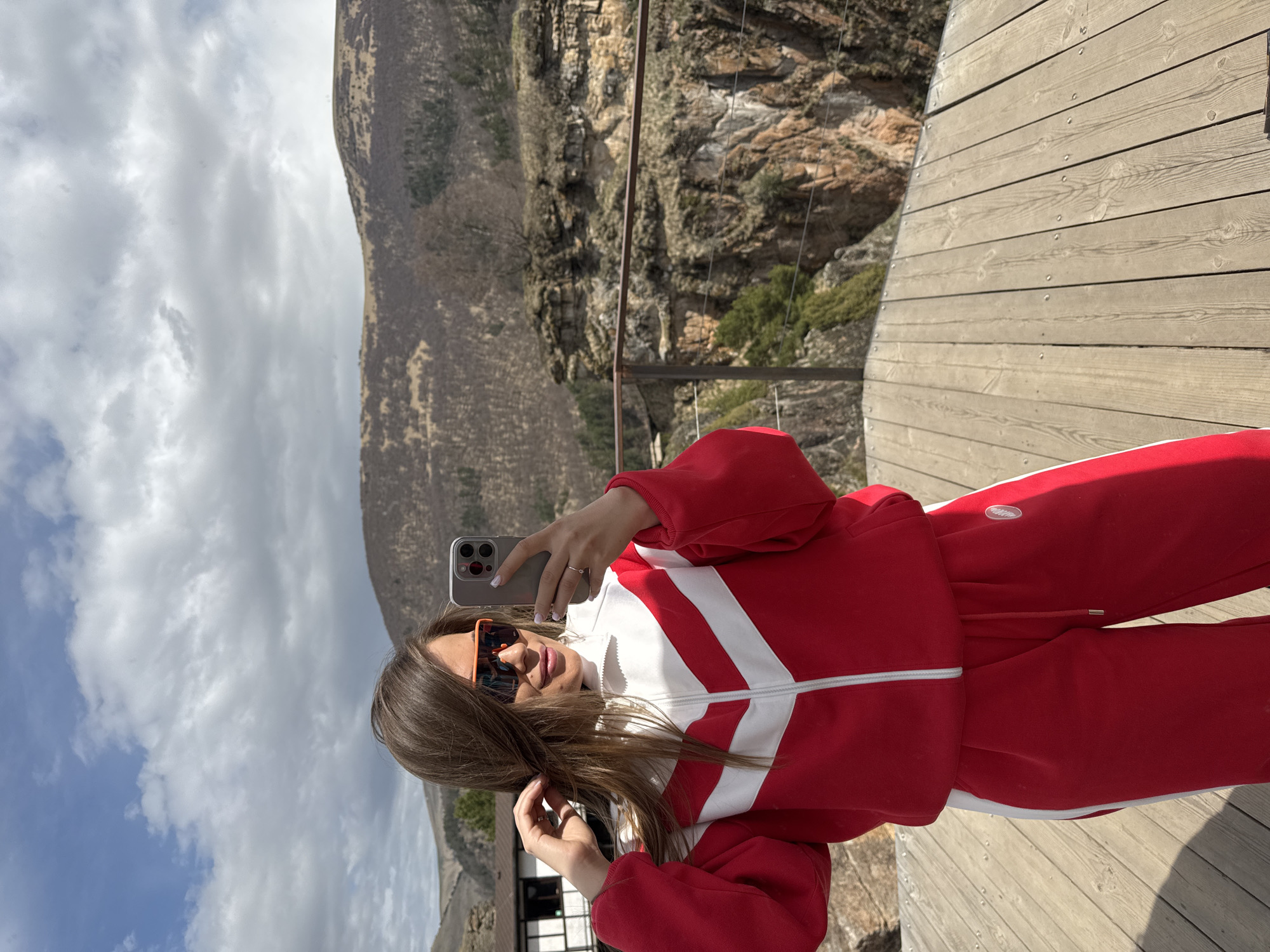A woman takes a selfie outdoors on a wooden platform. She wears a red tracksuit with white accents and orange-tinted sunglasses. She is holding a smartphone up to take the picture. The background shows a mountain landscape with a cloudy sky. The location is in the city of Unknown, Russia.