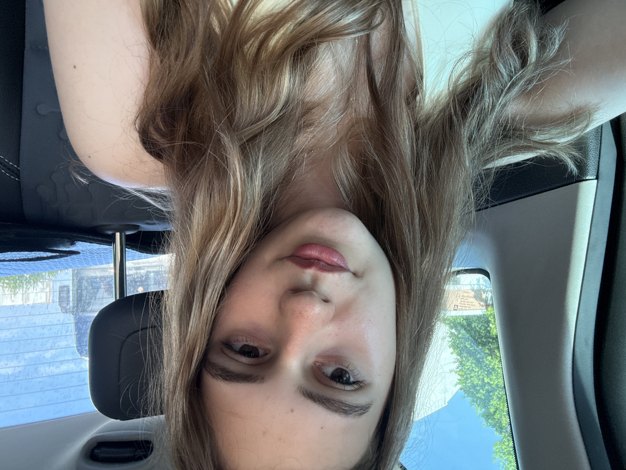 A young female, appearing to be in her late teens or early twenties, is depicted from the chest up, positioned in the rear passenger seat of a vehicle in the city of Plovdiv, Bulgaria. She has fair skin, dark brown eyes looking slightly downward and to her left, and long, wavy, light brown hair with blonde highlights that cascade over her shoulders and chest. Her lips are tinted with a reddish-pink hue, and minor skin texture and small freckles are visible on her nose and cheeks. She wears a light-colored top, possibly cream or white, with a broad strap visible over her right shoulder.
The vehicle's interior features dark-toned upholstery, visible as a headrest behind her left shoulder and parts of the window frame. Through the left side window, a clear blue sky, vibrant green tree foliage, and a light-colored building are visible. The rear window displays reflections of another vehicle, possibly a bus or truck, and distant urban infrastructure, overlaid with a textured pattern on the glass. The scene is illuminated by bright daylight, indicating clear weather conditions.