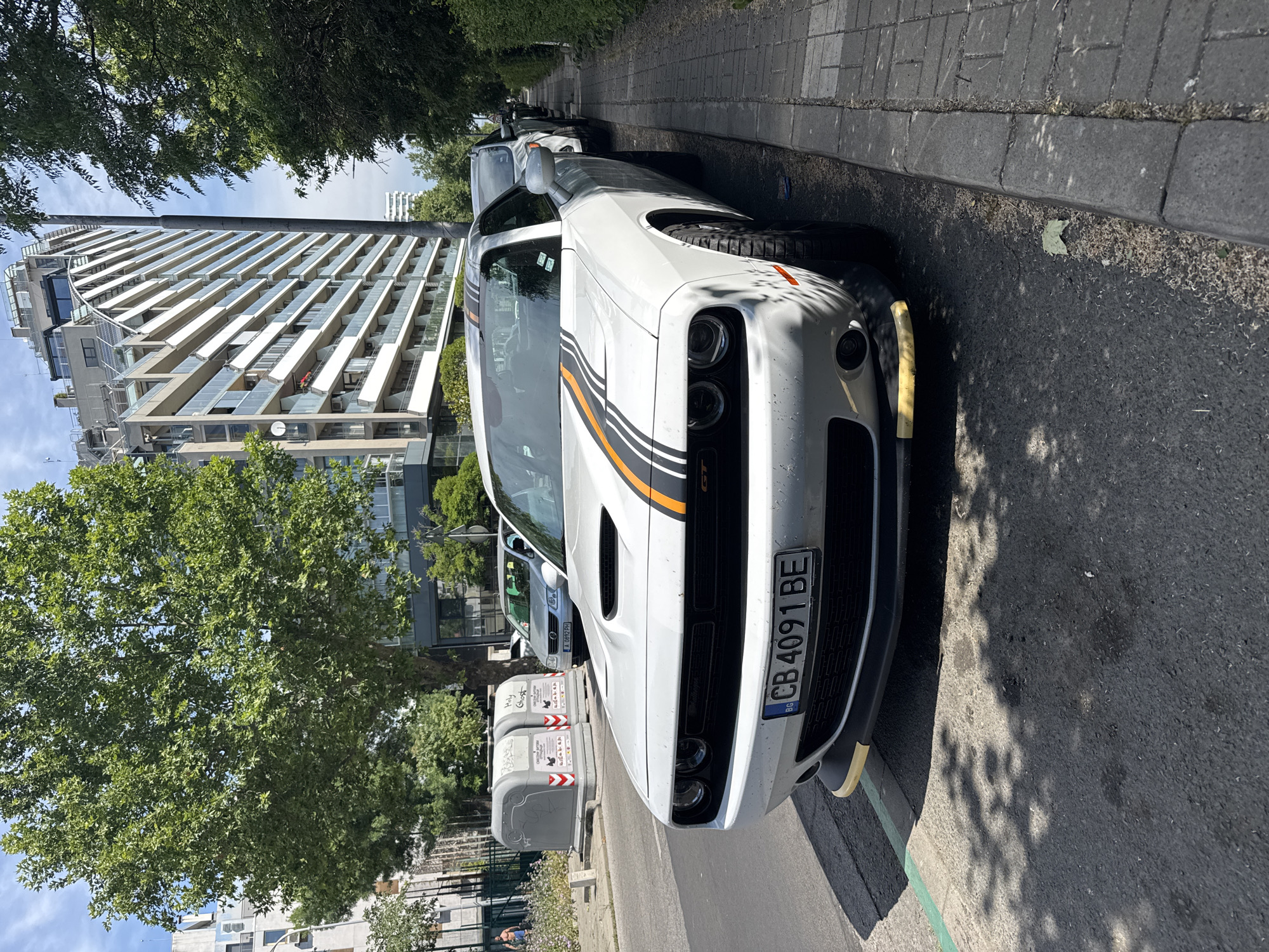 A white Dodge Challenger with orange and black racing stripes is parked on a street in Burgas, Bulgaria. The car has a Bulgarian license plate reading "CB 4091 BE". In the background, there is a modern apartment building with balconies and large windows, as well as a large leafy tree. Two grey refuse bins are also visible on the sidewalk. The scene is illuminated by natural sunlight, casting shadows on the street. The perspective is low-angle, focusing on the front of the car.
