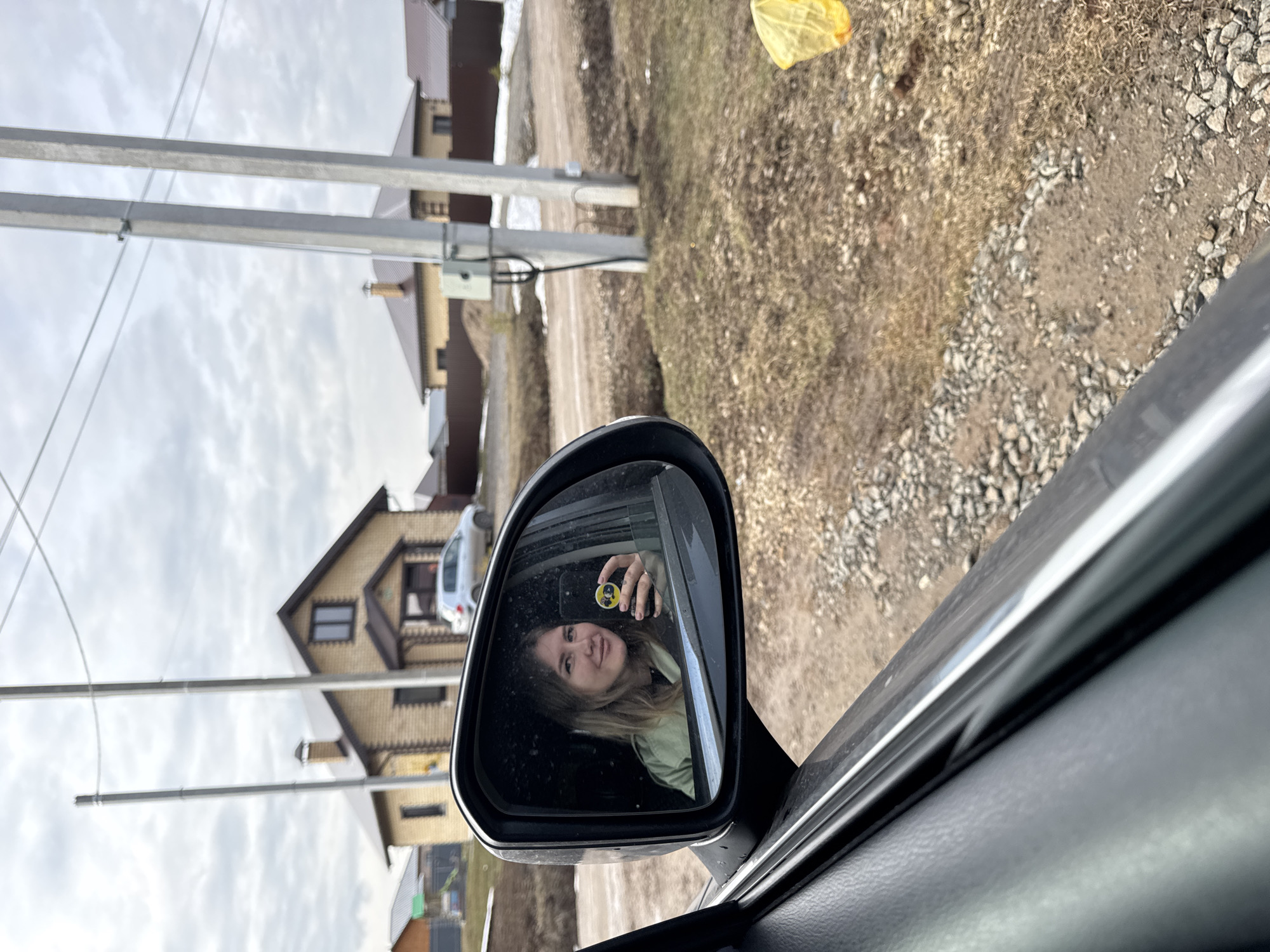 A woman is visible in the side mirror of a car. She is fair-skinned with blonde hair and is holding a small, round object with a bee emblem on it. The car appears to be parked on a dirt road in a residential area. In the background, houses and utility poles are visible. The sky is overcast. The location is identified as the city of Gornyi, Dvurechenskoe S/P, Russia.