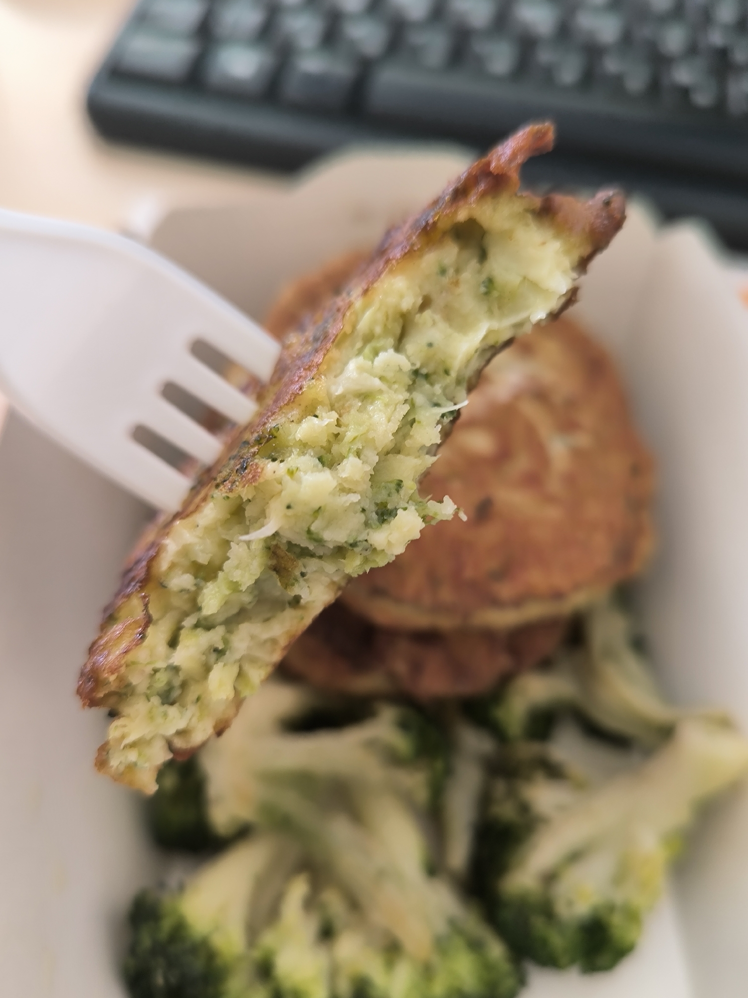 A close-up shot shows a fork picking up a bite from a cooked broccoli fritter. The fritter is golden brown on the outside and appears to be made with mashed vegetables, likely potatoes and broccoli. In the background, several more fritters are stacked in a white paper container, and a bed of steamed broccoli florets is visible below. A blurred keyboard is visible in the upper left corner of the image. The scene appears to be indoors, possibly at a desk or a cafe, and there is no indication of its location in Novosibirsk, Russia.