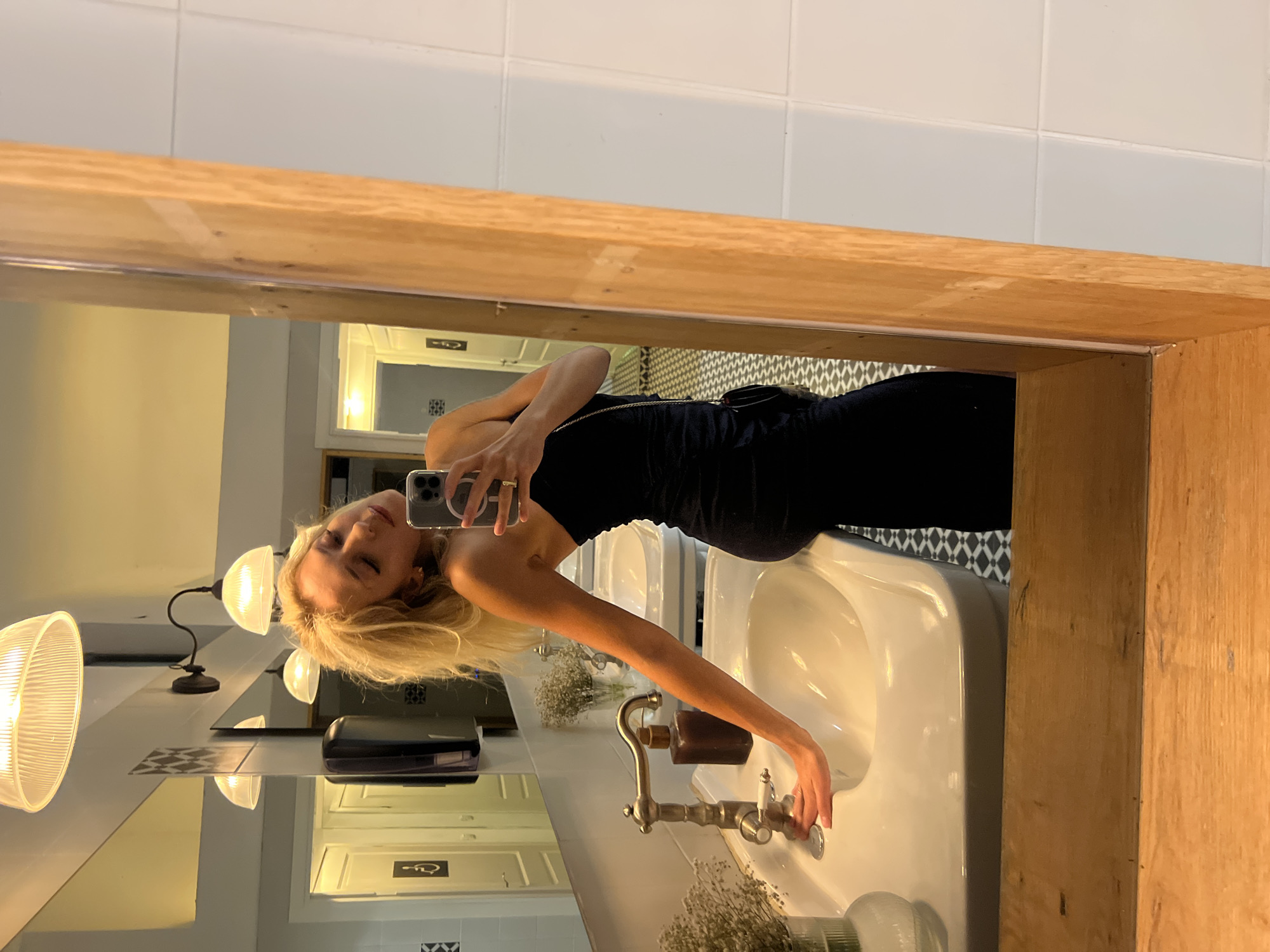 In Beit Hanan, Israel, a young blonde woman is taking a mirror selfie in a dimly lit restroom. She is wearing a strapless black dress and has a small purse with a chain strap slung over her shoulder. Her right hand rests on a white sink, and she is holding a phone in her left hand to capture the image. The background shows a tiled wall, a mirror reflecting the woman and the restroom, and a wooden frame. The sink area includes a faucet and a dispenser.