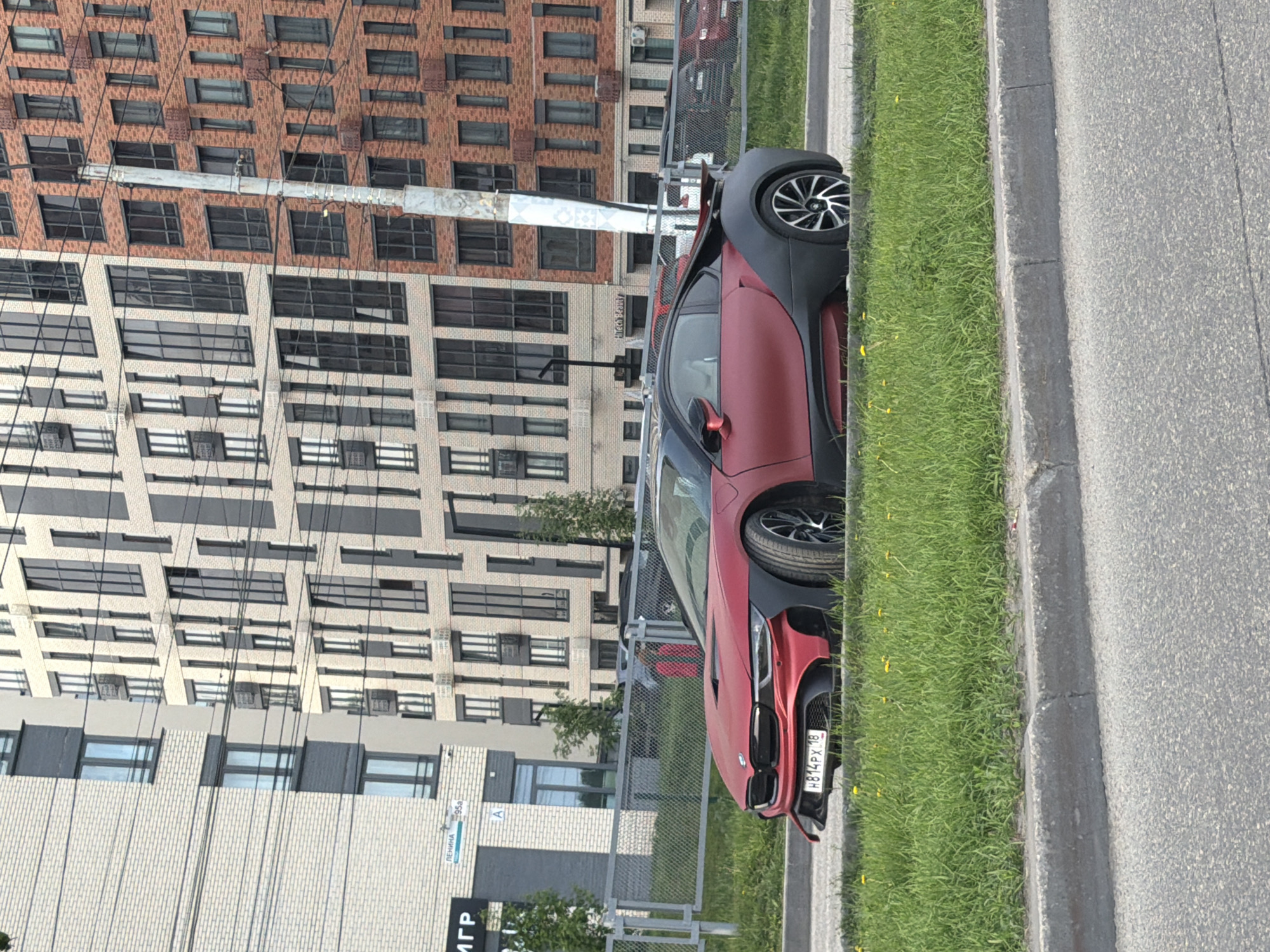 The image depicts a red and black BMW i8 parked on the side of a road, next to a strip of grass and a curb. In the background, there are apartment buildings with multiple windows. Based on the sign visible in the lower-left corner indicating "Ленина 95а" (Lenina 95a), and the license plate indicating Russia, this photo seems to be taken in the city of Izhevsk, Russia. The license plate number appears to be H814PX 18. No people are visible. The car is the main subject. The environment appears to be urban.