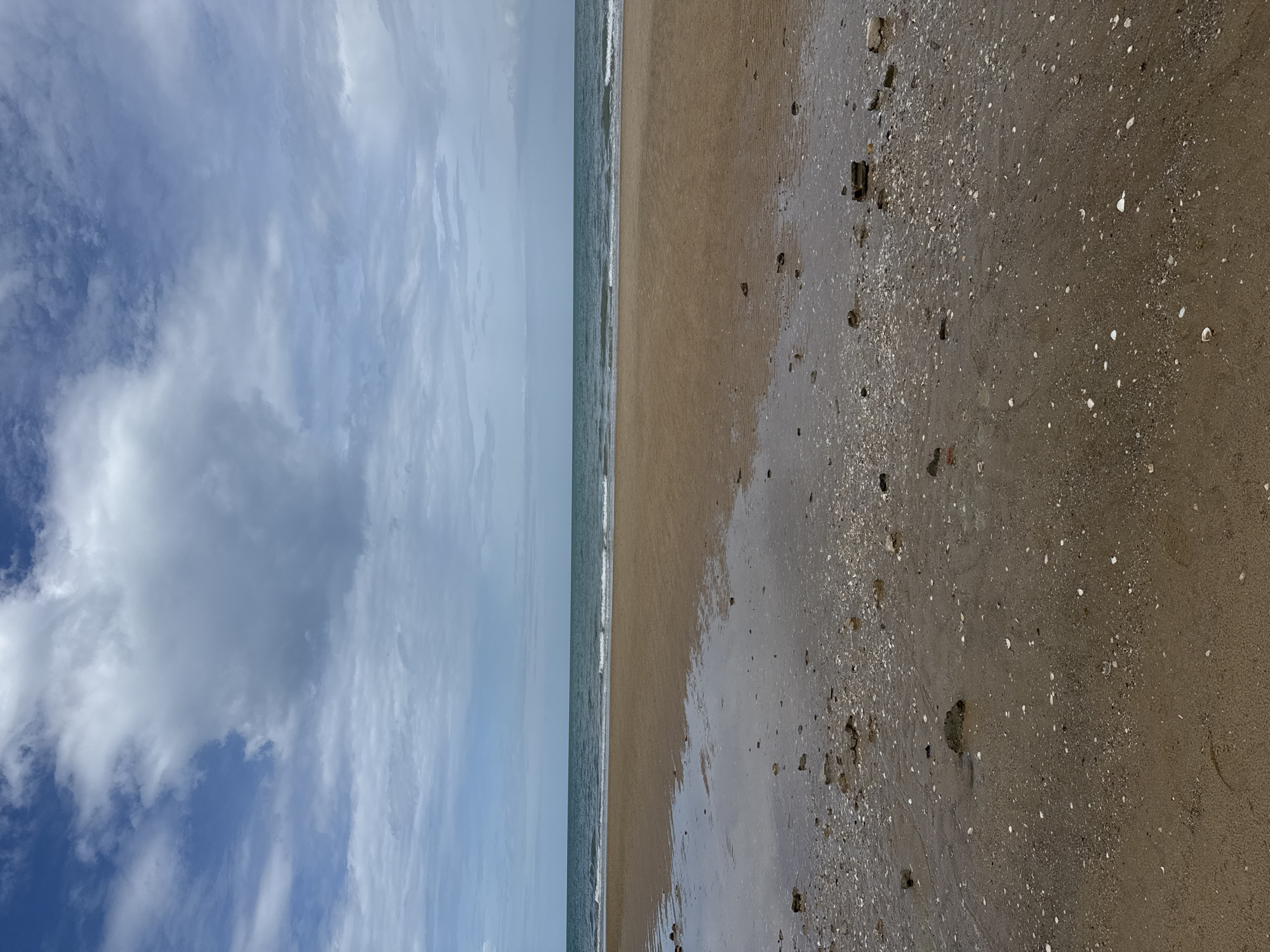 The image shows a wide, sandy beach with calm, blue-green ocean waves gently lapping the shore. The sky is filled with a mix of white and gray clouds, suggesting an overcast or partly cloudy day. Scattered across the wet sand in the foreground are small shells and pebbles. The scene depicts a natural coastal environment. This location is identified as Ban Bang Ket, Thailand.