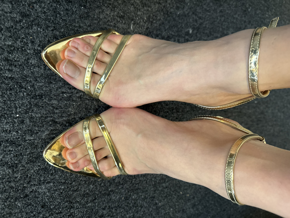 The image presents a top-down, close-up view of two human feet positioned on a dark gray, short-pile carpet. This scene is located in Arcueil, France. Each foot wears an identical gold-colored, open-toed, high-heeled sandal. The sandals feature two parallel, reflective gold straps across the forefoot, exposing the toes, and a slender gold ankle strap with a visible buckle mechanism on the left foot's strap. The individual's skin tone appears light. The toenails are natural in color and unpolished. The carpet surface shows minor lint or debris. No other individuals, actions, or significant objects are visible within the frame.