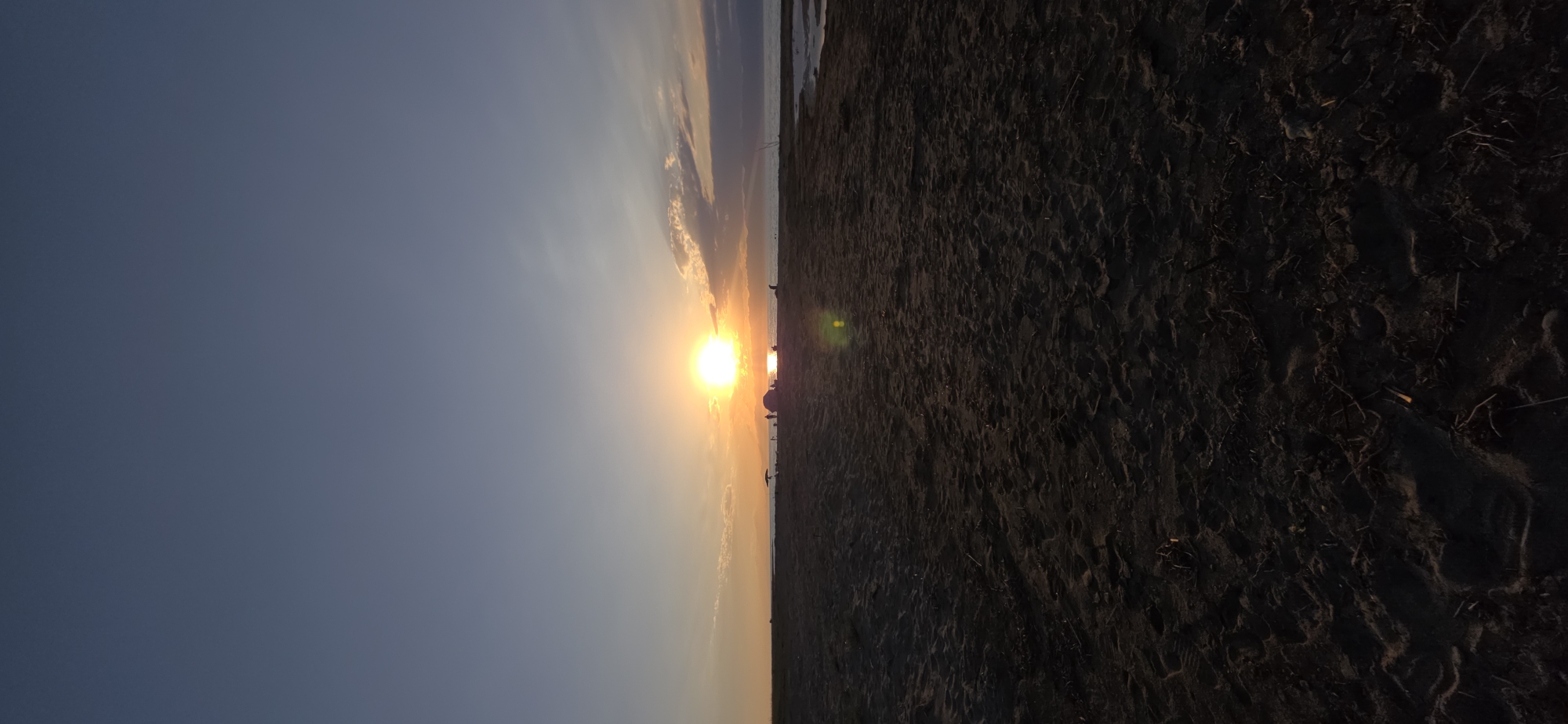 The image captures a serene sunset over a beach in Boz Beshik, Kyrgyzstan. The sky is a gradient of deep blue at the top transitioning to lighter hues near the horizon, with soft clouds tinged orange by the setting sun. The sun itself is a bright, radiant orb positioned just above the distant horizon, casting a warm glow across the scene.
In the middle ground, a stretch of dark sand forms the beach. Scattered across it are several small groups of people, appearing as silhouetted figures against the bright sky. Some individuals seem to be standing, while others might be seated or engaged in activities near what appears to be a beach umbrella and perhaps a tent. The sand itself is textured, showing footprints and small debris, suggesting recent activity. To the right, a calm body of water is visible, reflecting the colors of the sunset. The foreground is dominated by the dark, textured sand, drawing the viewer's eye towards the illuminated horizon.