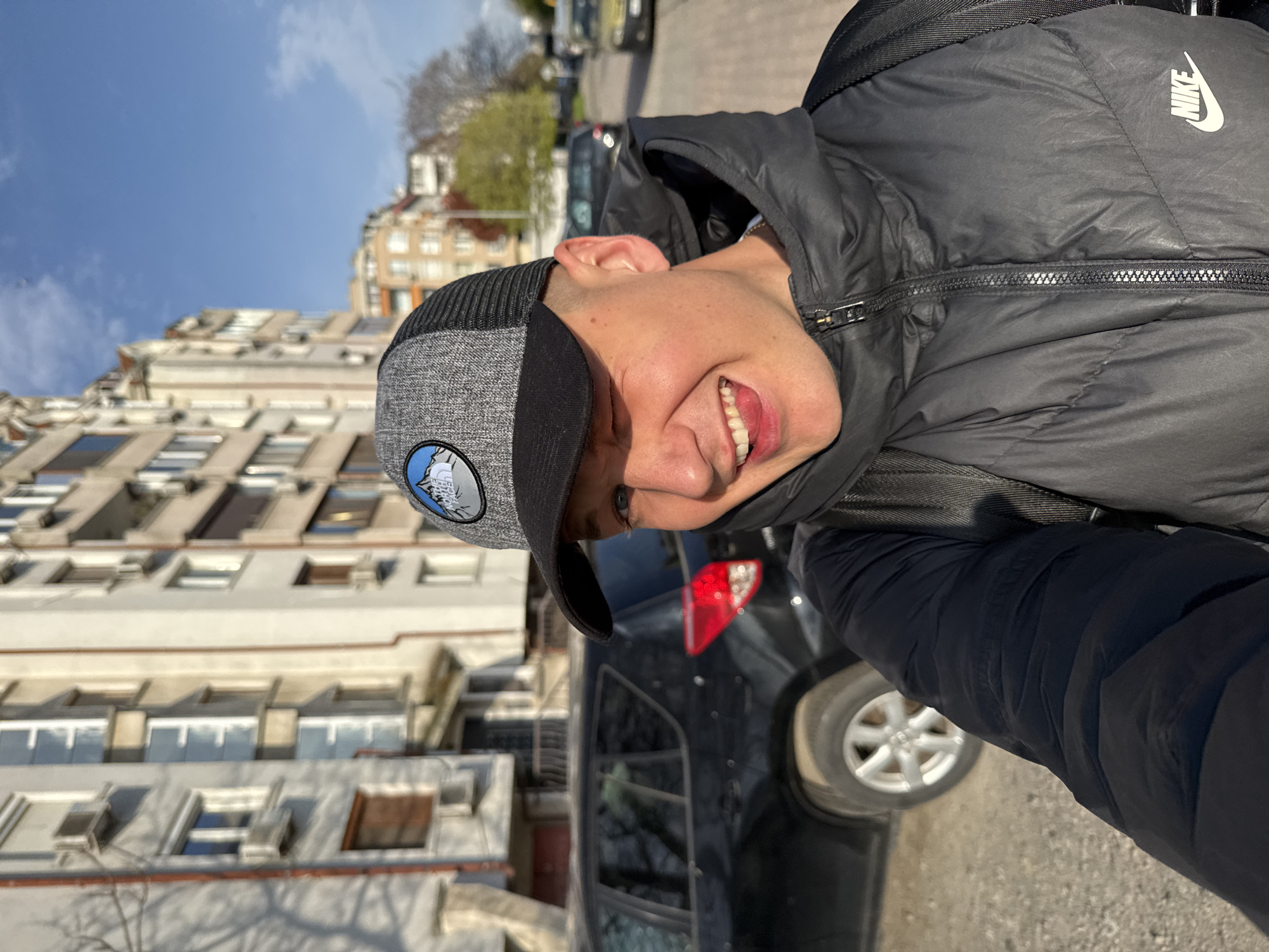 A young man in a black Nike puffer jacket and grey baseball cap stands on a street in Burgas, Bulgaria. He smiles at the camera. Behind him is a black SUV, multi-story buildings, and a partially visible tree against a blue sky. The image is slightly rotated counter-clockwise.