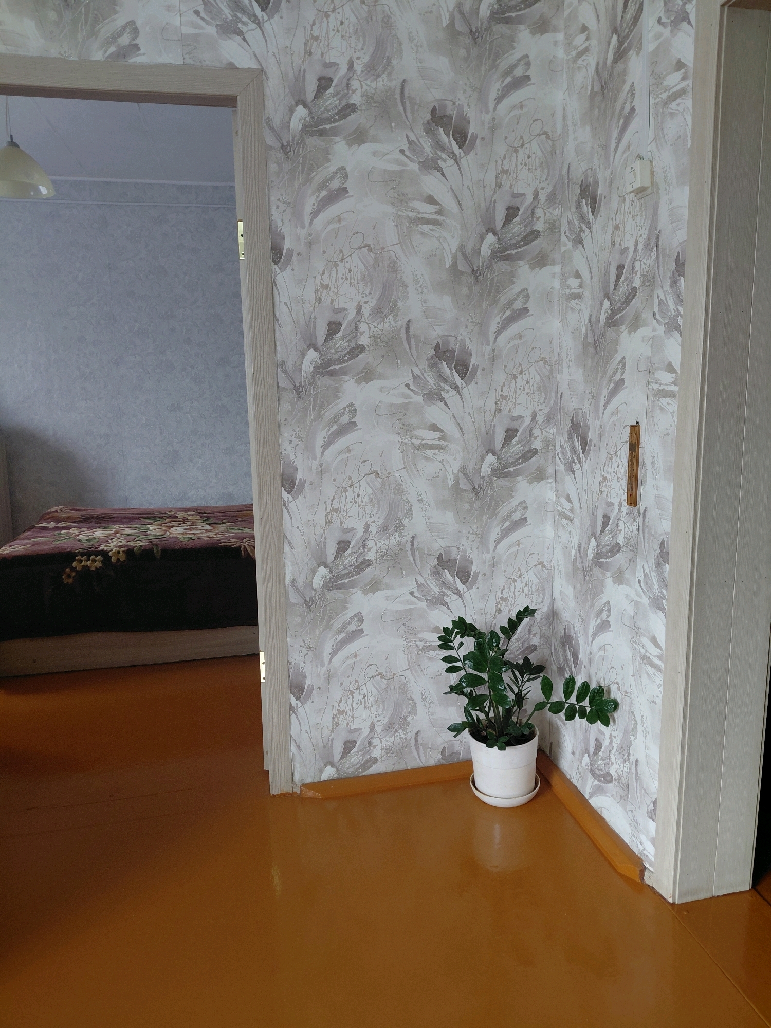 The image depicts an interior view of a room in an apartment building in Malinovka, Russia, likely taken from a hallway. The primary focus is on the corner of a room where a doorway opens into another space. The wallpaper in the corner features a distressed, floral pattern in shades of grey, beige, and white. A white ceramic pot containing a green potted plant is situated on the light brown wooden floor, directly in the corner. To the right of the plant, a pale wooden door frame is visible. In the background, through the open doorway, a bed with a dark burgundy blanket adorned with a floral pattern is partially visible. The floor throughout the visible area is a glossy, light brown color. A white ceiling light fixture with a pale yellow shade is faintly visible in the upper left portion of the image, through the doorway. A wooden thermometer hangs on the wall to the right of the potted plant. The scene appears to be a domestic interior, showing a residential living space.