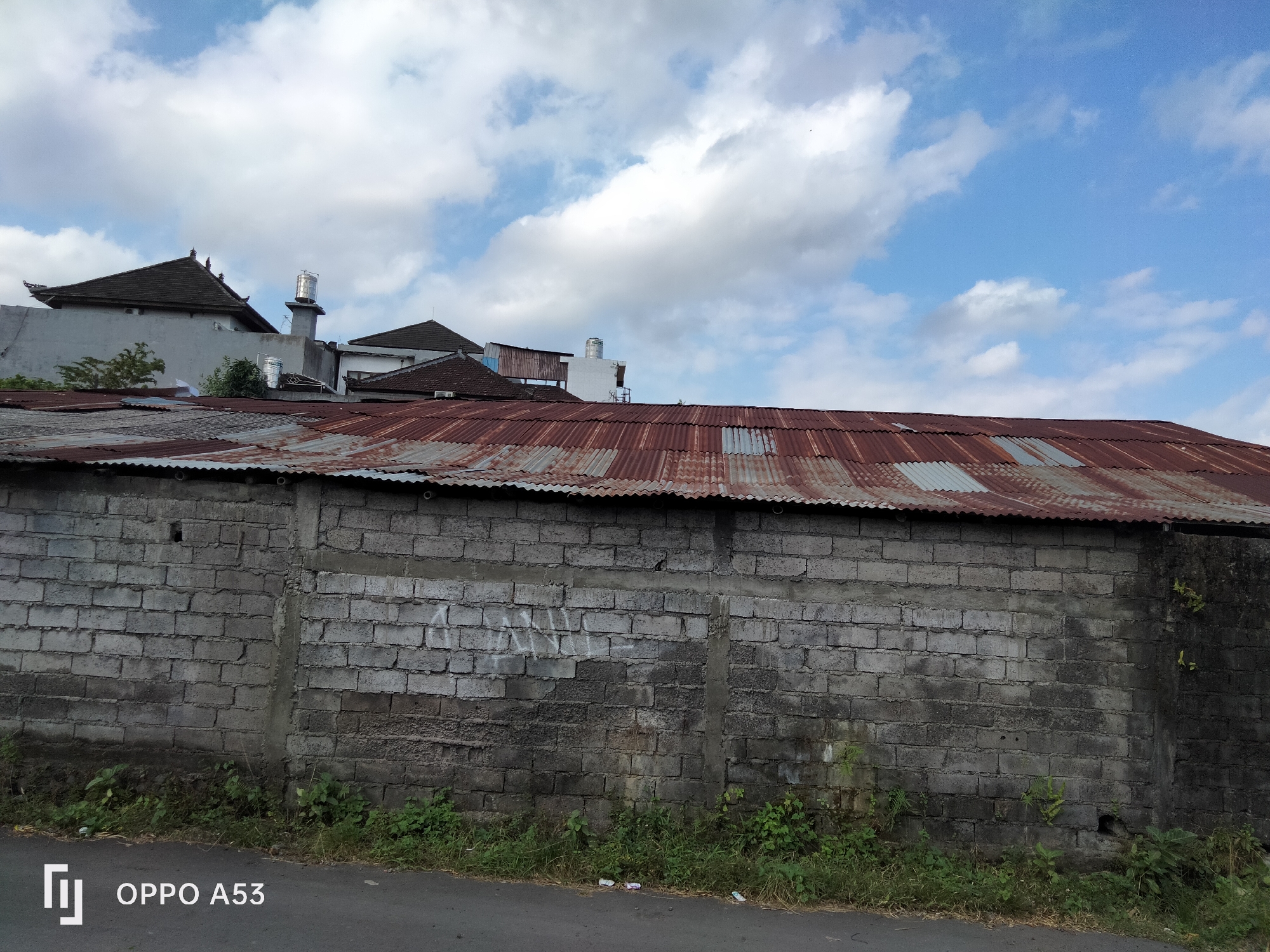 The image depicts a street scene in Denpasar, Indonesia. A rough, grey brick wall runs horizontally across the mid-ground, topped by a weathered, corrugated metal roof. Beyond the wall, several buildings are visible, including structures with traditional Balinese-style roofs and a silver water tank on a pedestal. The sky above is a vibrant blue with scattered white clouds. Vegetation, including grass and small plants, grows along the base of the wall and at the edge of the asphalt road in the foreground. The road itself shows some debris. The image is branded with "OPPO A53" in the lower left corner.