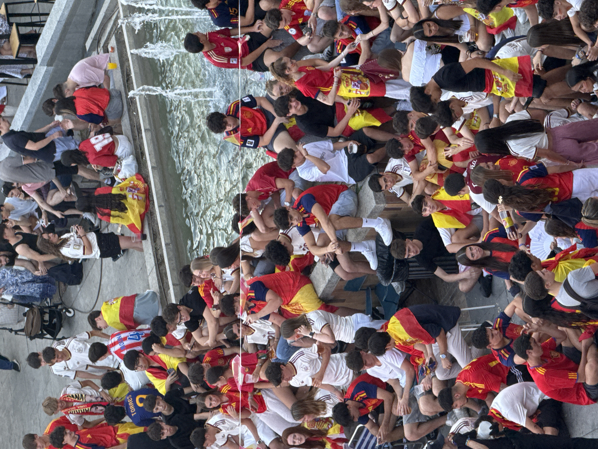 The image depicts a large group of people, primarily young adults and teenagers, gathered outdoors, seated on the edge of a fountain and surrounding pavement. Many are wearing red and yellow attire, suggesting support for a sports team or national event, possibly related to Spain given the presence of Spanish flags draped over shoulders and held by individuals.
The environment appears to be an urban public space. The fountain's water is visible, with jets creating a spray. The ground is paved, and there are stone ledges which people are using as seating. In the background, elements of urban infrastructure can be seen, including what looks like a stroller, seating structures, and possibly scaffolding or construction elements. Some individuals are holding drinks, and the overall atmosphere suggests a casual gathering or event. The clothing styles are contemporary.
While the image contextually suggests a Spanish setting, there is no explicit visual information within the frame that definitively identifies the location as Las Rozas de Madrid, Spain. The scene is a snapshot of a public gathering, with a focus on the individuals and their shared activity.