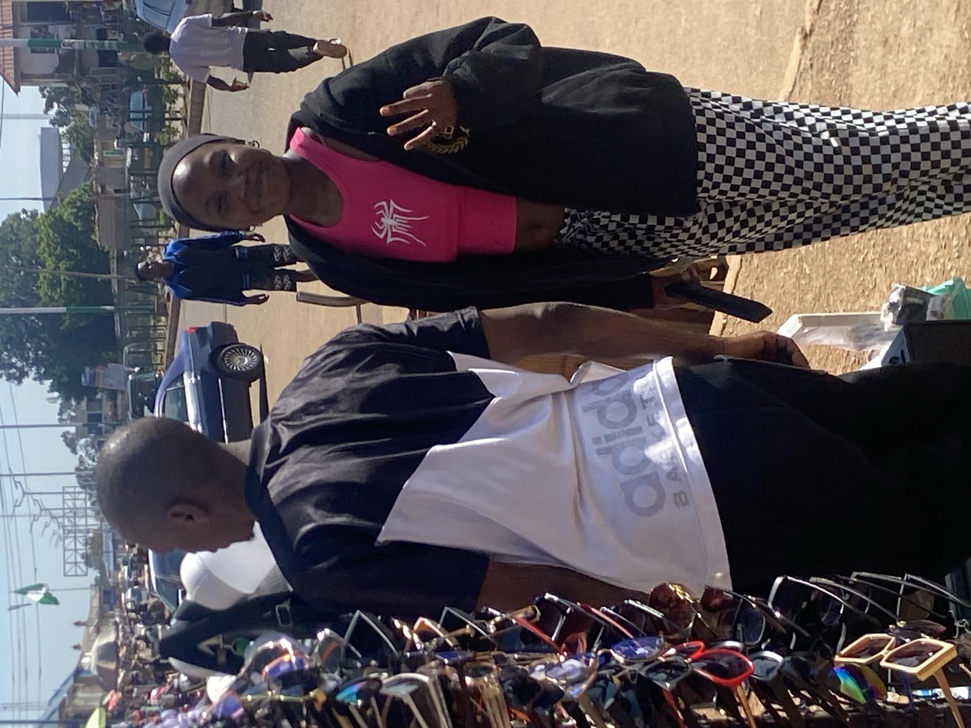 An outdoor street scene in Bukuru, Nigeria, features two individuals prominently. In the foreground, a person wearing a black hoodie over a pink tank top with a spiderweb graphic, and black and white checkered pants, smiles at the camera. They are also wearing a black headscarf and a ring on their right hand, with fingers held in a "peace" sign. To the left, a person in a black and white t-shirt and black pants stands with their back to the camera, facing a display of sunglasses. In the background, several cars, buildings, and people are visible, along with a Nigerian flag. The setting appears to be a market or busy street.