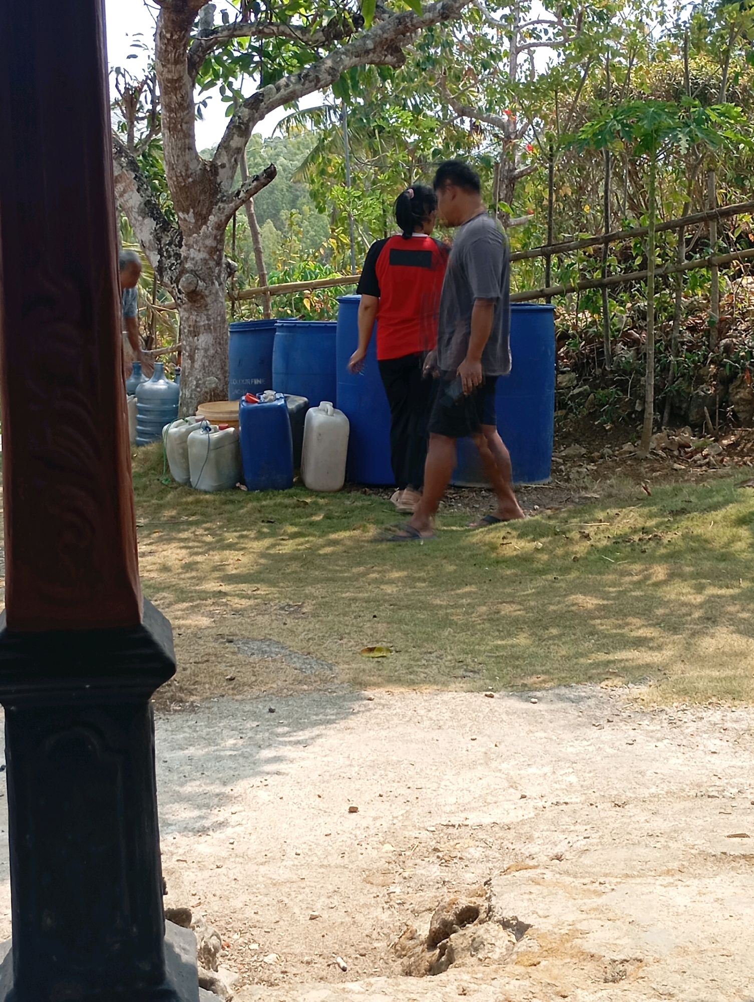 In the city of Ketepung, Indonesia, a scene unfolds outdoors with a focus on a collection of large blue barrels, several smaller white and grey plastic containers, and water jugs. Two individuals are present. One person, wearing a red shirt with black sleeves and black pants, stands facing away from the camera. To their right, a person in a grey t-shirt and black shorts is also facing away, holding a dark object. An older person, partially visible, is tending to some of the water jugs in the background. The environment consists of dry, dusty ground, a grassy area with scattered leaves, and lush green vegetation, including trees with visible bark patterns and bamboo fencing. The scene suggests a functional, possibly agricultural or water storage area, bathed in bright sunlight.