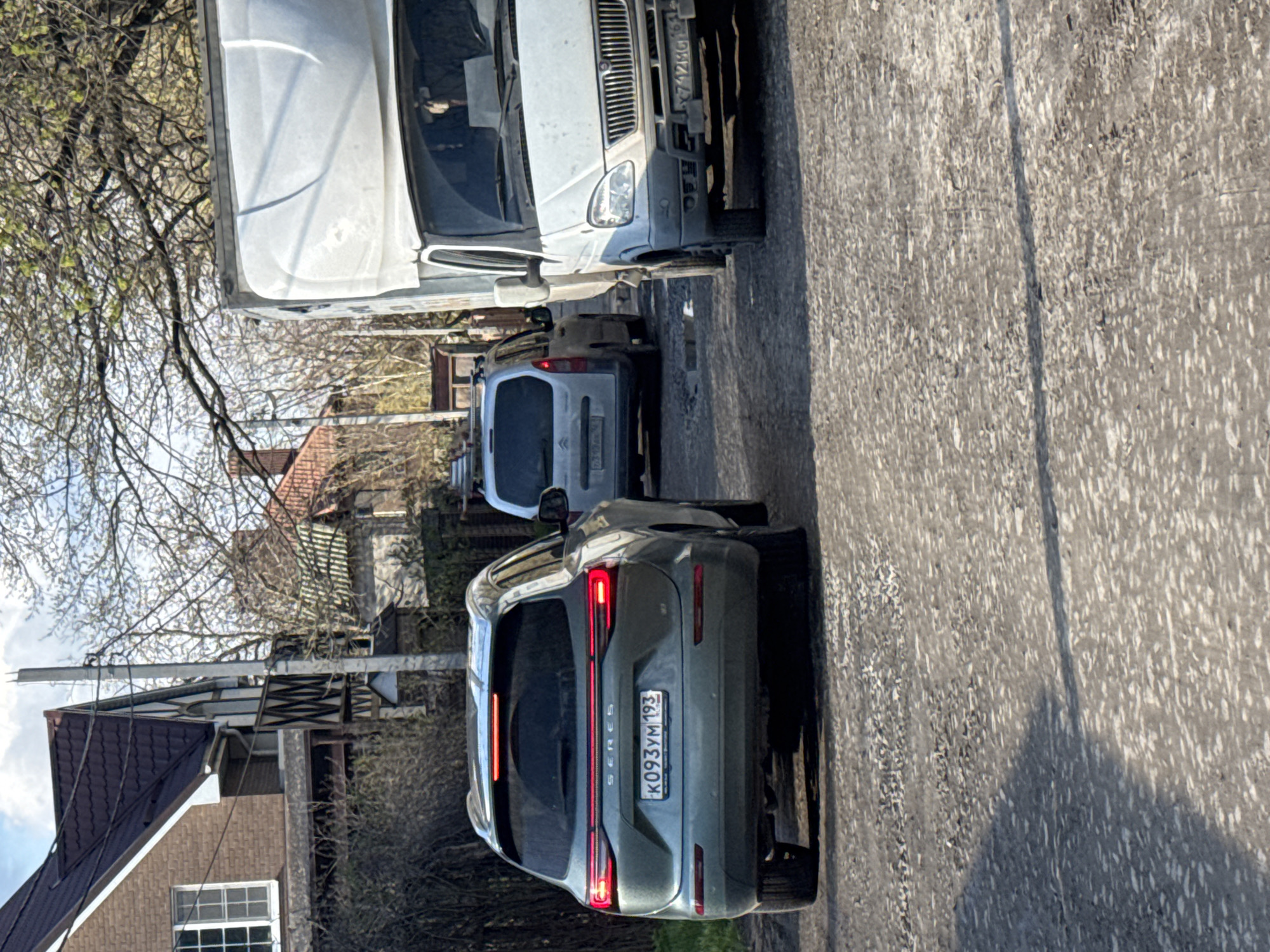 The image depicts a street scene in Rostov-on-Don, Russia, featuring several parked vehicles. A light grey or green-colored SUV with license plate "K093YM 193" is prominently positioned in the foreground. A white car, possibly a van or small truck, is parked in front of the SUV. A light-colored compact car is visible between the two. The road surface is asphalt and appears worn. Buildings, including a house with a brown brick facade and a dark roof, are visible in the background on the left side of the image. Bare trees are present, indicating it is likely early spring or late autumn. Overhead power lines are also visible.