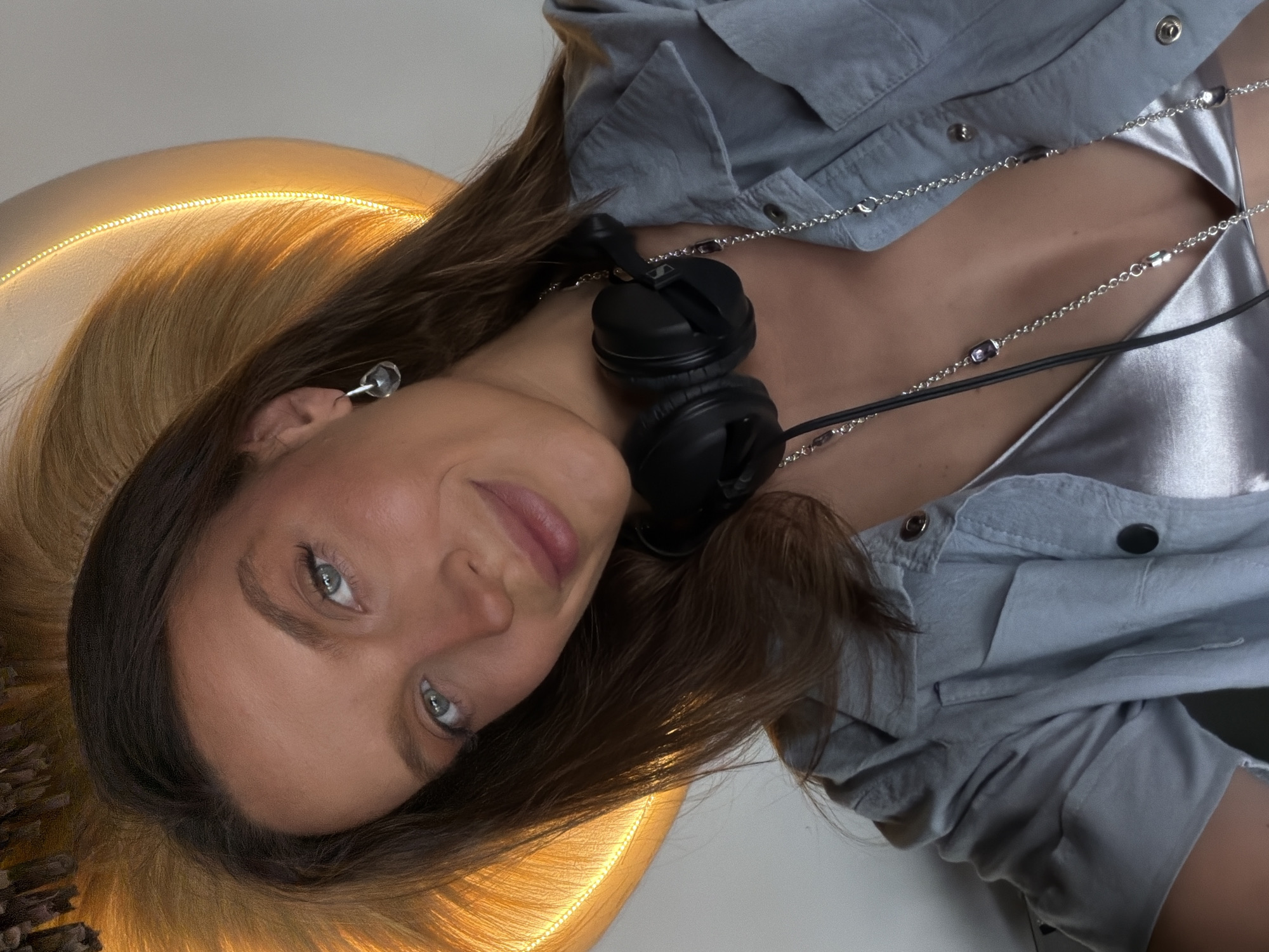 A medium shot photograph shows a young woman with long, dark brown hair and light blue eyes, smiling at the camera. She is wearing headphones around her neck, a grey collared shirt over a shimmering silver top, and a silver necklace with purple beads. She has a small earring in her left ear. The background features a large, round object with a bright yellow light emanating from its edge, casting a warm glow. The overall scene appears to be indoors, possibly in a studio or a creatively lit space. The image does not contain any specific elements that would suggest a location in Drobnići, Montenegro.