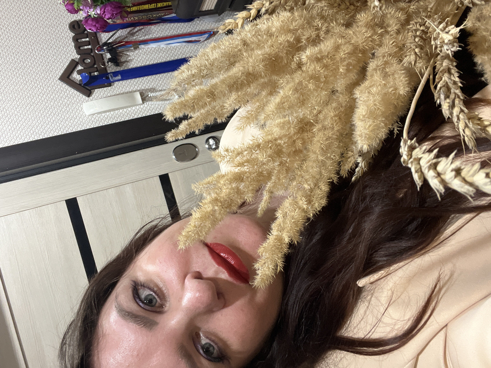 A close-up, slightly rotated image shows a woman with dark brown hair and red lipstick. Her face is prominent, with visible details of her eyes and skin. She is positioned next to a bouquet of dried wheat and pampas grass. Behind her, a wall is visible. On the wall is a wall decor piece of a house marker/arrow sign with purple flower decorations. Also on the wall is a white intercom. The location is Noviy, Russia.