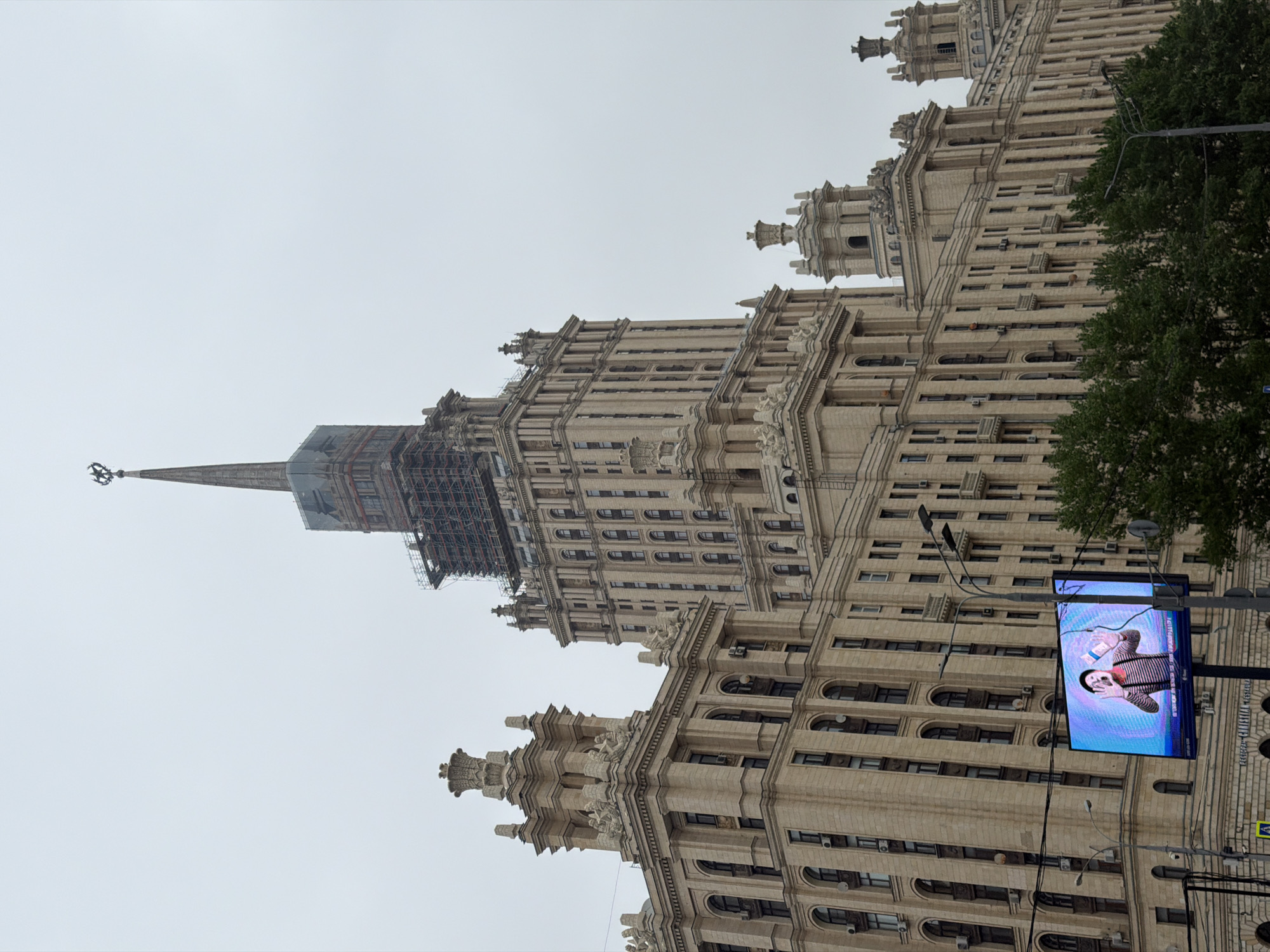 The image depicts the Ministry of Foreign Affairs building, one of the Seven Sisters skyscrapers in Moscow, Russia. The building is captured from a low-angle perspective, showcasing its intricate architectural details and towering height. The sky is overcast and gray, muting the colors of the building. A large digital billboard is positioned in the lower right corner, displaying an image of a mime artist. A tree is visible behind the billboard. The spire of the building is partially covered in scaffolding.