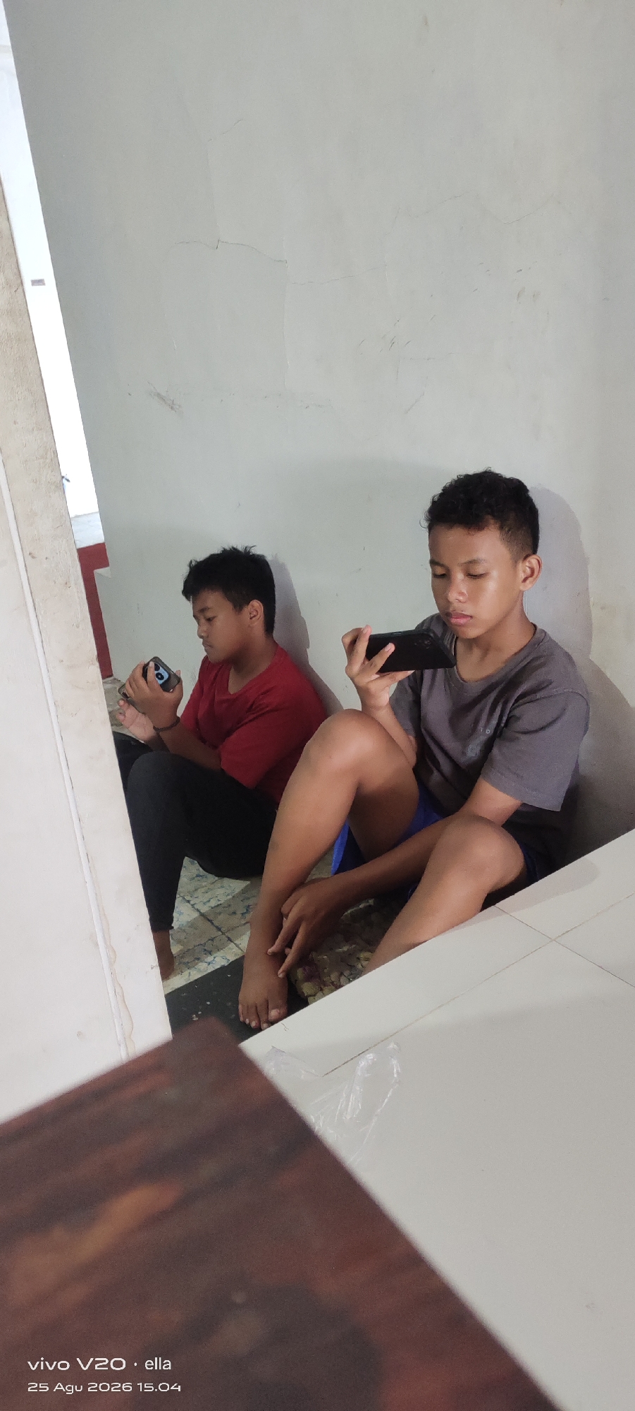 Two young boys are sitting on the floor next to a wall and a staircase, both looking at their phones. The boy on the left, wearing a red shirt and black pants, holds a phone in his left hand. The boy on the right, wearing a grey t-shirt and blue shorts, holds a phone in his right hand. Their feet are visible, with the boy on the right barefoot. The environment appears to be indoors, possibly a house or building with a tiled floor and a white, somewhat textured wall. The lighting suggests it is daytime. The bottom of the image shows a partial view of a wooden surface and text indicating "vivo V20 · ella" and a date and time stamp of "25 Agu 2026 15.04". The location is identified as Simo, Indonesia.