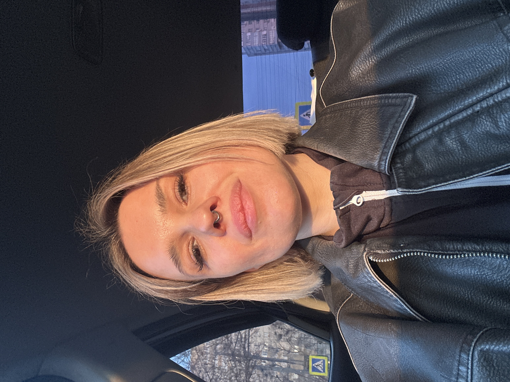 The image shows a female individual seated within the passenger compartment of a vehicle in Rostov-on-Don, Russia. The woman, with shoulder-length blonde hair and a septum piercing, wears a black faux leather jacket with white contrast stitching over a dark hooded sweatshirt. She is looking directly at the camera, indicating a self-portrait. Strong, low-angle sunlight illuminates the right side of her face and jacket. Through the vehicle's left rear window, an urban street scene is visible, including bare trees, a multi-story building, and a yellow square pedestrian crossing sign with a blue triangle and white pedestrian figure. A taller, light-colored multi-story building is partially visible through the right window against a clear blue sky. The lighting conditions suggest either late afternoon or early morning.