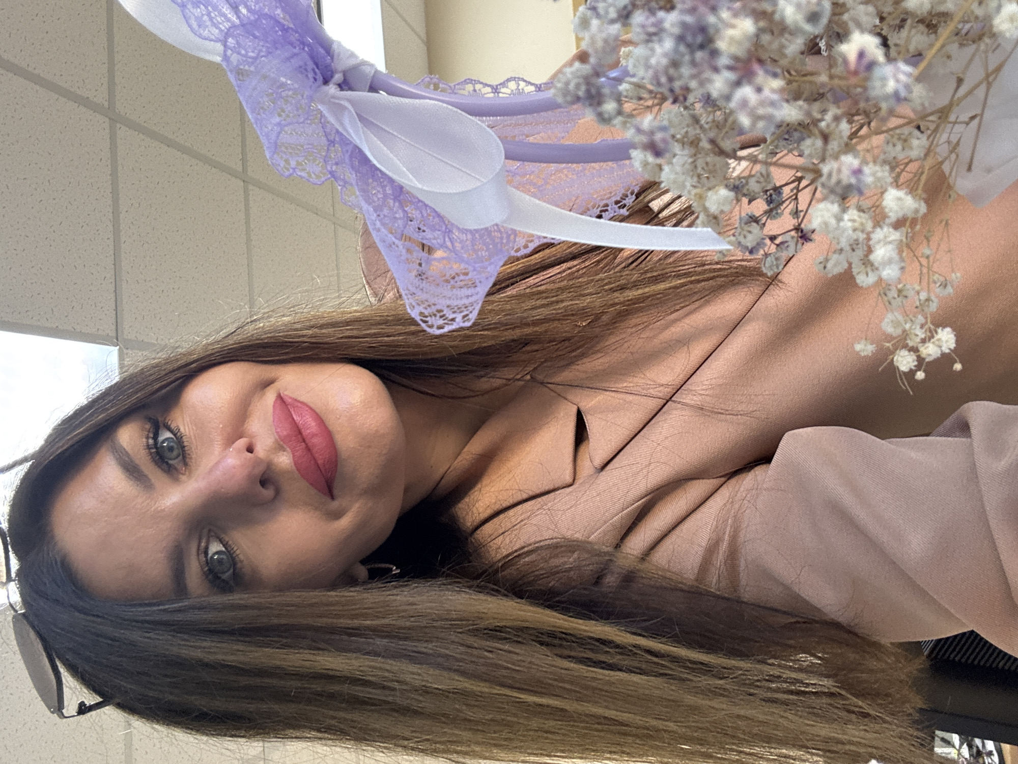 A medium shot, chest-up, selfie of a woman with long, brown hair and bright blue eyes, wearing a light brown blazer and pink lipstick. She is holding a bouquet of dried baby's breath flowers and a decorative item adorned with light purple lace and a white ribbon. The background appears to be an indoor office space with a suspended ceiling. The woman is positioned in the foreground, with the bouquet and decorative item partially obscuring her right side. The lighting is bright, suggesting an artificial light source or daylight from a window. No specific location in Chaikovsky, Russia, is identifiable from the image.