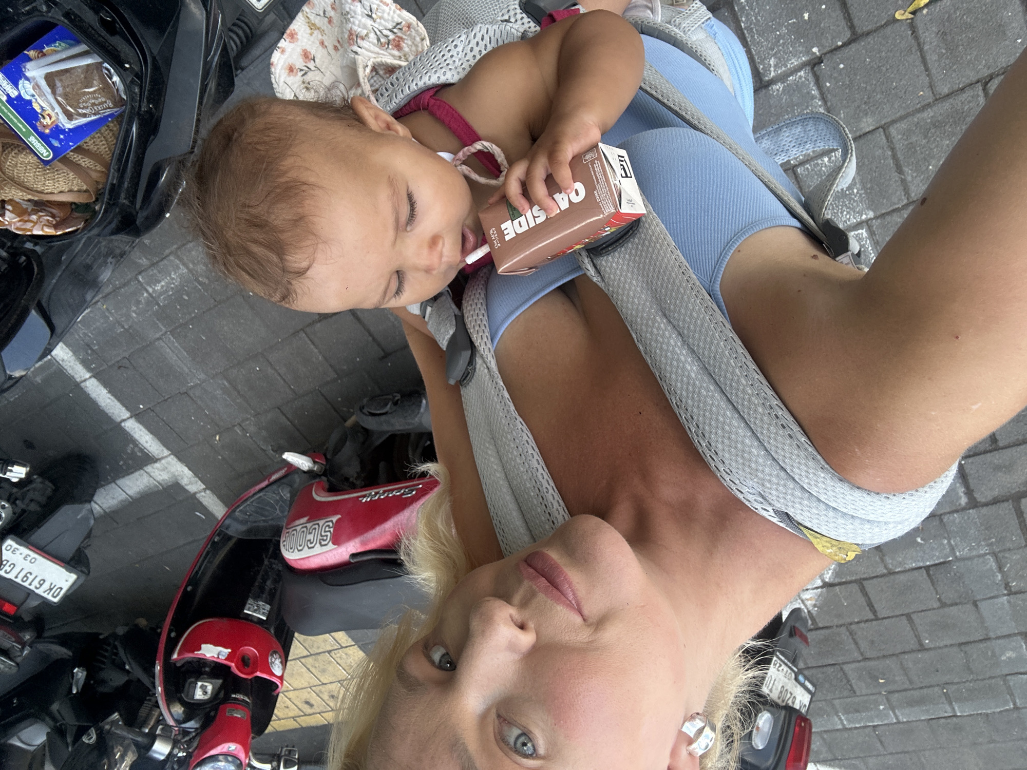 In North Kuta, Indonesia, a woman is taking a selfie with a baby in a carrier. The woman has blonde hair, blue eyes, and is wearing a blue tank top and large hoop earrings. The baby, with dark curly hair and brown eyes, is being held in a gray baby carrier and is drinking from a small carton of OATSIDE chocolate milk with a straw. The background shows a paved area with parked scooters, one of which has a license plate starting with "DK 6191". A woven bag and a box of Nestlé snacks are visible in the storage compartment of a black scooter in the foreground.