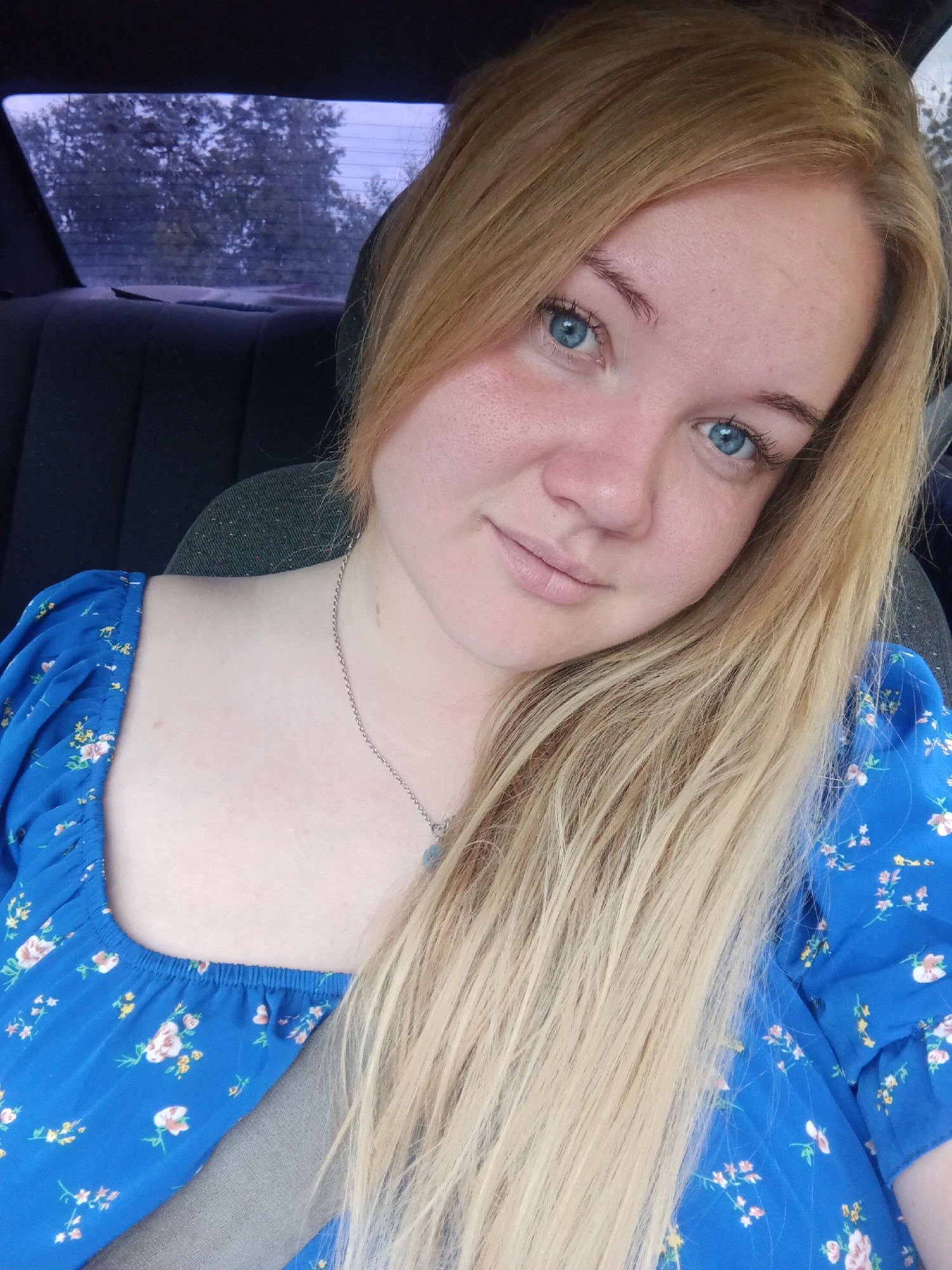 A young Caucasian woman with fair skin, blue eyes, and long blonde hair is pictured from the chest up, seated in the passenger seat of a vehicle. She is wearing a blue dress with a floral pattern and a silver necklace. The background shows the rear window of the car, through which trees are visible against an overcast sky, suggesting an outdoor setting, possibly in a suburban or rural area of Chaikovsky, Russia. The lighting is natural, indicating daytime.