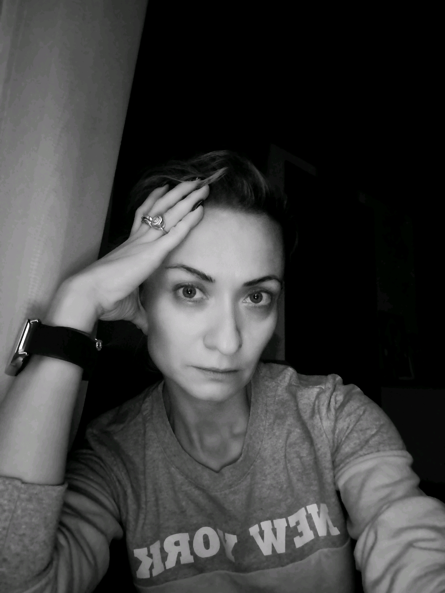 A black and white, close-up, waist-up, selfie of a woman is shown. She is resting her left arm and hand on her forehead, with her fingers splayed and touching her temple. She is wearing a grey long-sleeved shirt with the words "NEW YORK" printed across the chest in white block letters. A watch with a black band is visible on her left wrist. She has short, dark hair and her gaze is directed at the camera. The background is dark with a light-colored vertical object on the left side of the frame, which appears to be a wall or curtain. The woman is not in Moscow, Russia.
