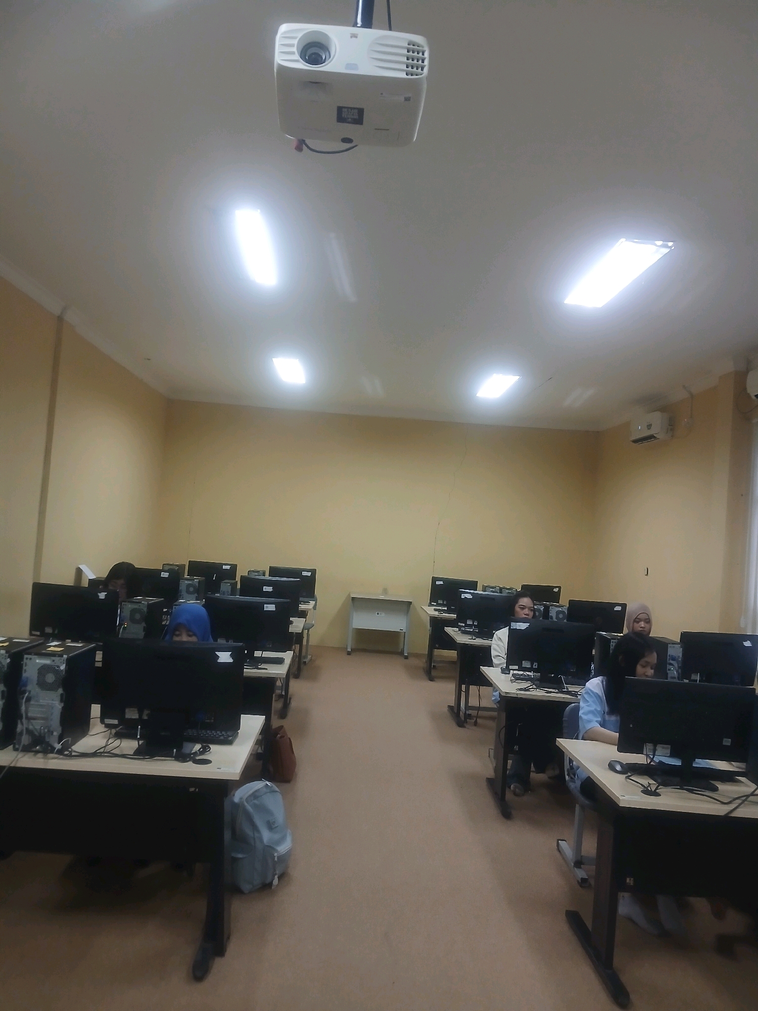The image depicts a computer lab, likely in an educational institution in Kedungrejo, Indonesia, as suggested by the presence of multiple computer workstations and students. A ceiling-mounted projector is visible in the upper portion of the frame. The room has cream-colored walls and a beige carpeted floor. Several students, mostly young women, are seated at desks, engaged with their computer screens. Some are wearing hijabs. Desks are arranged in rows, each equipped with a desktop computer, monitor, and keyboard. Bags and personal items are visible around the workstations. The lighting appears to be from overhead fluorescent fixtures. The overall impression is of a typical classroom setting focused on computer-based learning.
