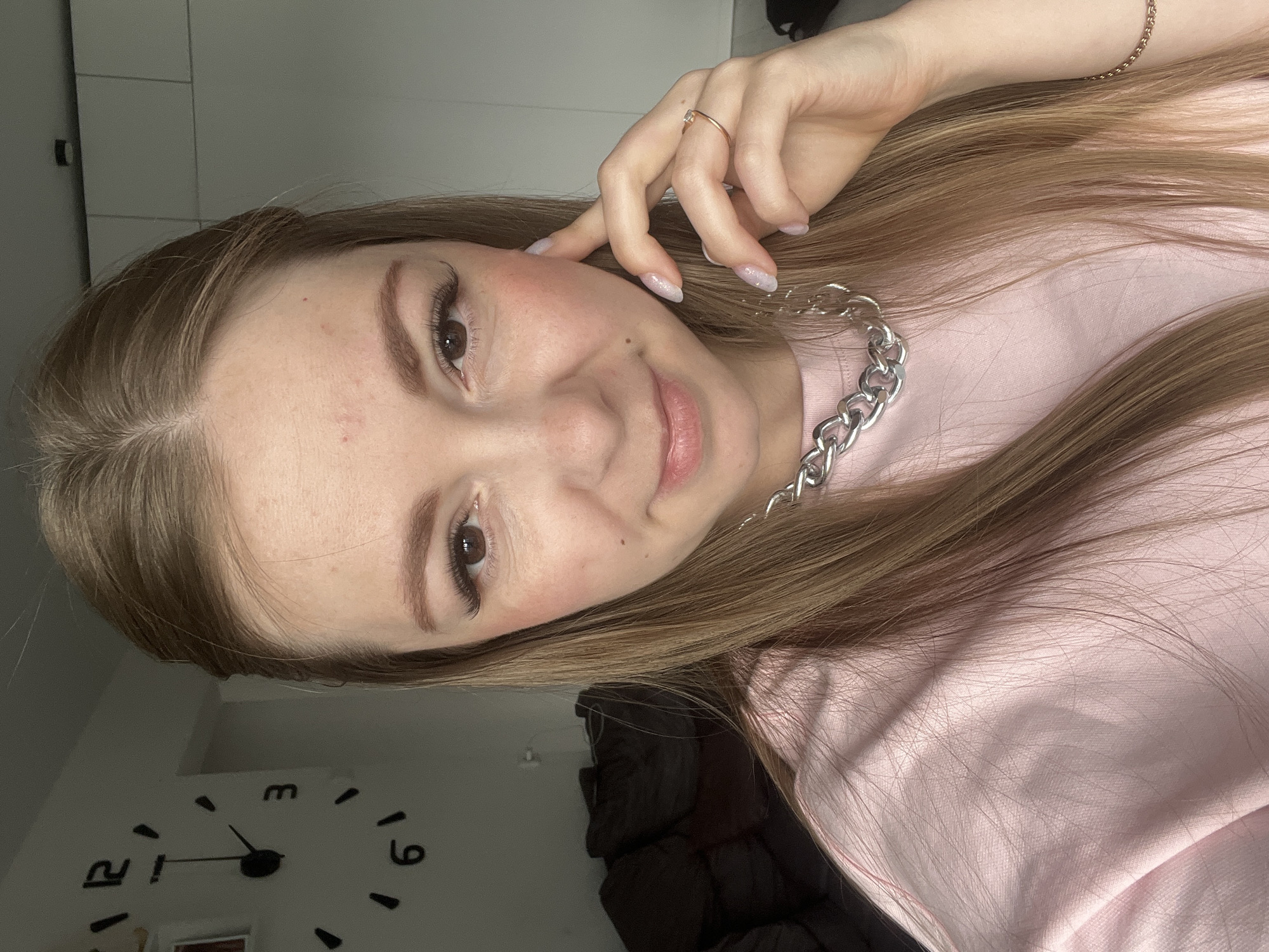 A young adult female is depicted in a close-up, chest-up portrait, likely a selfie, taken indoors in the city of Unknown, Unknown. She has long, straight, light brown hair parted slightly off-center to the left, falling over her shoulders. Her face, featuring brown eyes with visible long eyelashes, defined eyebrows, a small nose, and a slight smile with glossy lips, is turned directly towards the camera. She exhibits light blush on her cheeks and a few small blemishes on her forehead and right cheek. She wears a plain, light pink crew-neck top and a prominent, thick, silver-toned chain necklace. Her left hand (viewer's right) is raised, with manicured, light-colored, glittery nails, and a simple gold-toned ring with a clear stone on her ring finger, lightly touching her left cheek/jawline. A thin, gold-toned chain bracelet is partially visible on her right wrist. The background on the left features a white wall with a large, black-numbered wall clock, indicating approximately 12 o'clock. A dark brown sofa is visible out of focus behind her left shoulder. On the right, a light grey/white wall or cabinetry is present, with a small black rectangular fixture near the ceiling. The scene is brightly and evenly lit, suggesting natural light from an unseen window.