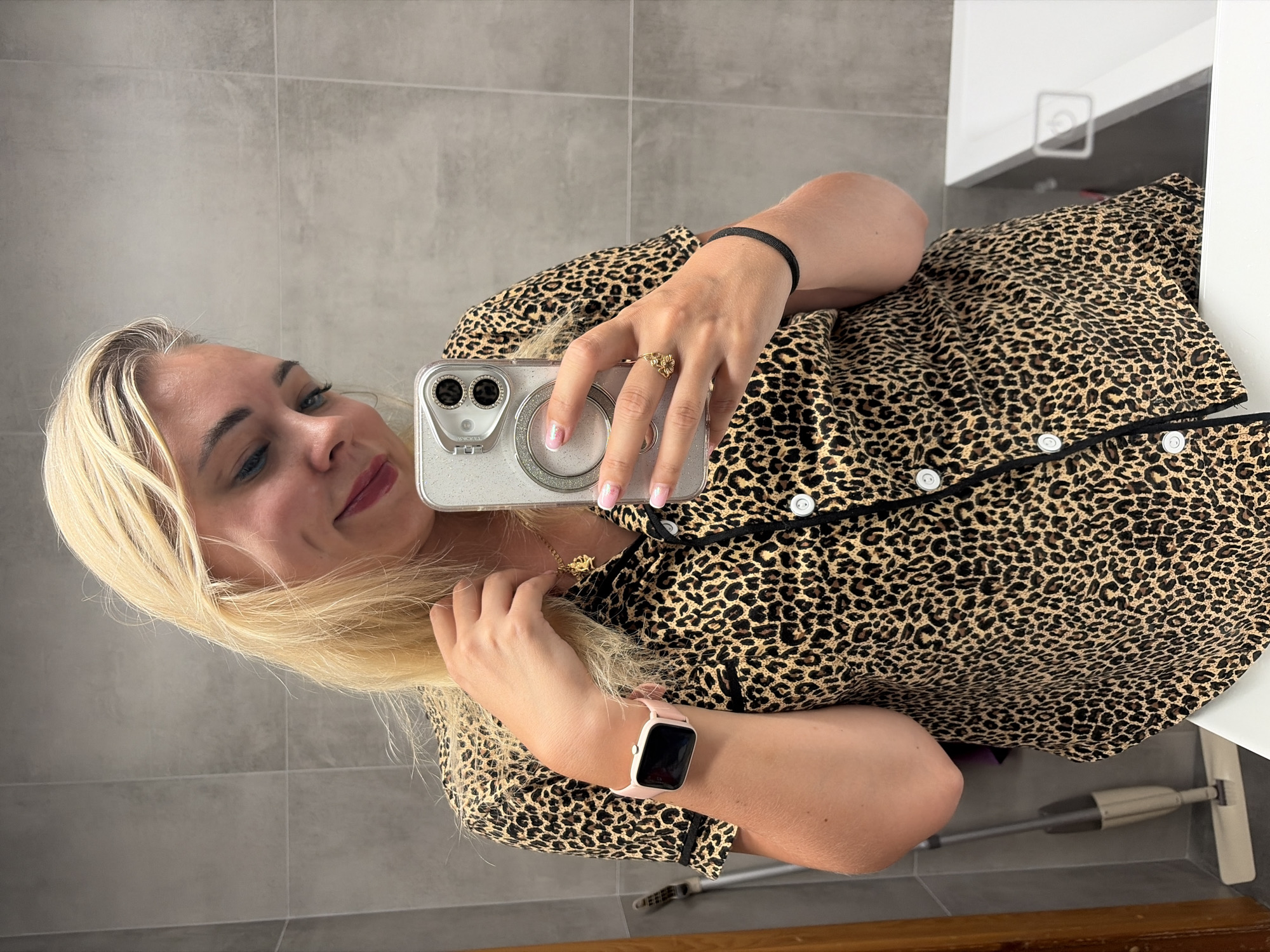 In the city of Chaikovsky, Russia, a blonde woman with dimples is taking a selfie in a bathroom. She is wearing a leopard-print pajama top and has a pink smartwatch on her left wrist. She is holding her hair with her right hand and has a gold ring on her index finger. The bathroom has gray tiled walls and a white bathtub. A mop is visible in the background.