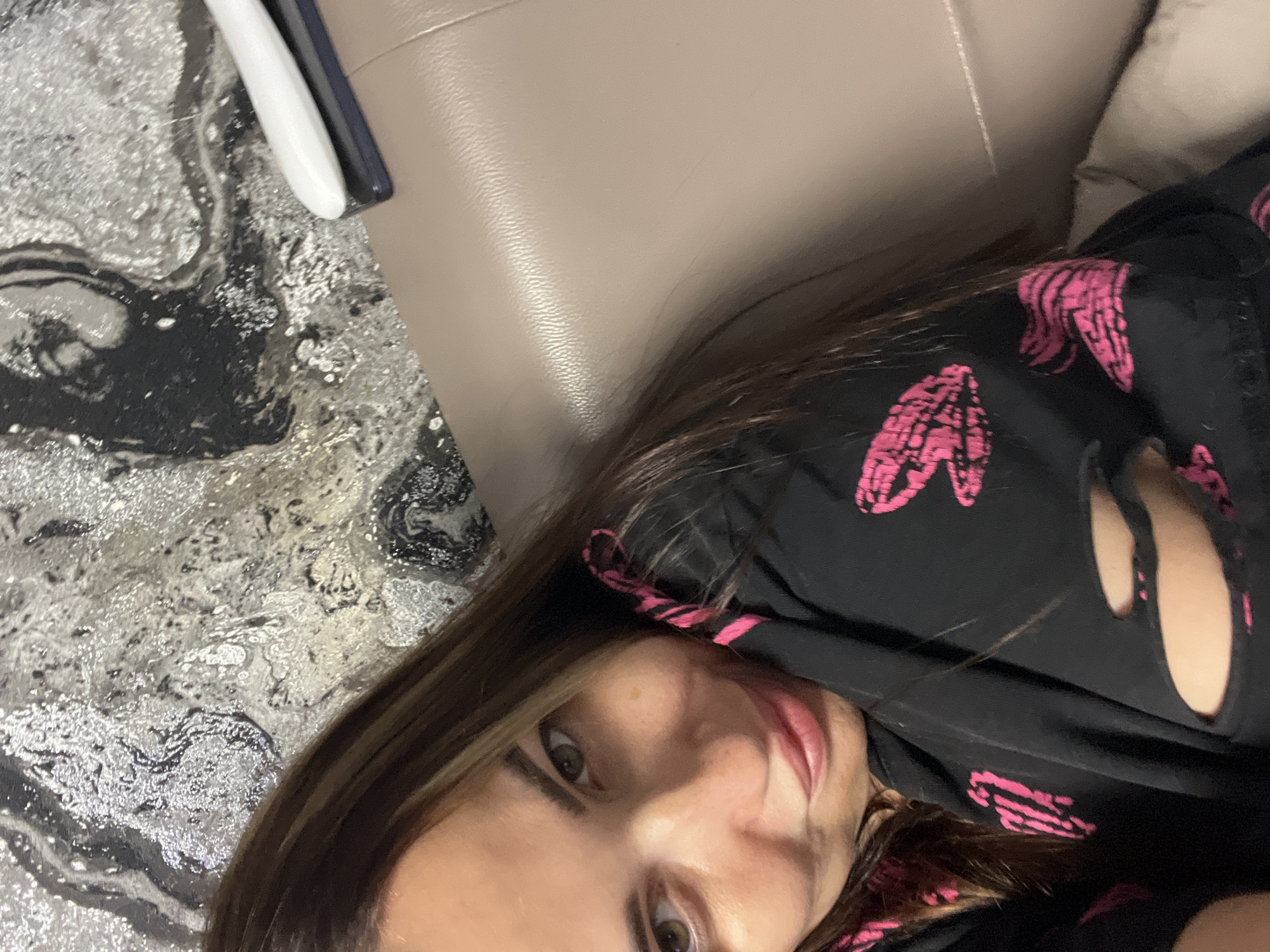 The media file is a close-up, rotated selfie of a woman. She has long, dark brown hair and light skin. Her eyes are a light green, and she is wearing dark eyeliner and light-colored eyeshadow. Her lips are full and appear to be painted with a pink or nude lipstick. She is wearing a black t-shirt with a repeating pink heart pattern. The t-shirt has cut-out details on the shoulders.
The background is partially visible and appears to be a mix of textures. To the left of the woman, there is a patterned surface that looks like a rug or a textured wall, with swirling black and silver patterns. To the right, there is a smooth, light brown surface, possibly a couch or furniture, with a slight sheen. In the upper left corner, a white object with a dark blue accent is visible, which might be a phone or a remote control.
The lighting suggests an indoor setting, possibly at night or in a dimly lit room, as there are some reflections on the smooth surface. The woman's expression is difficult to fully ascertain due to the angle and cropping, but her eyes are open and she seems to be looking directly at the camera. There is no visible text in the image. The overall mood is casual and personal.