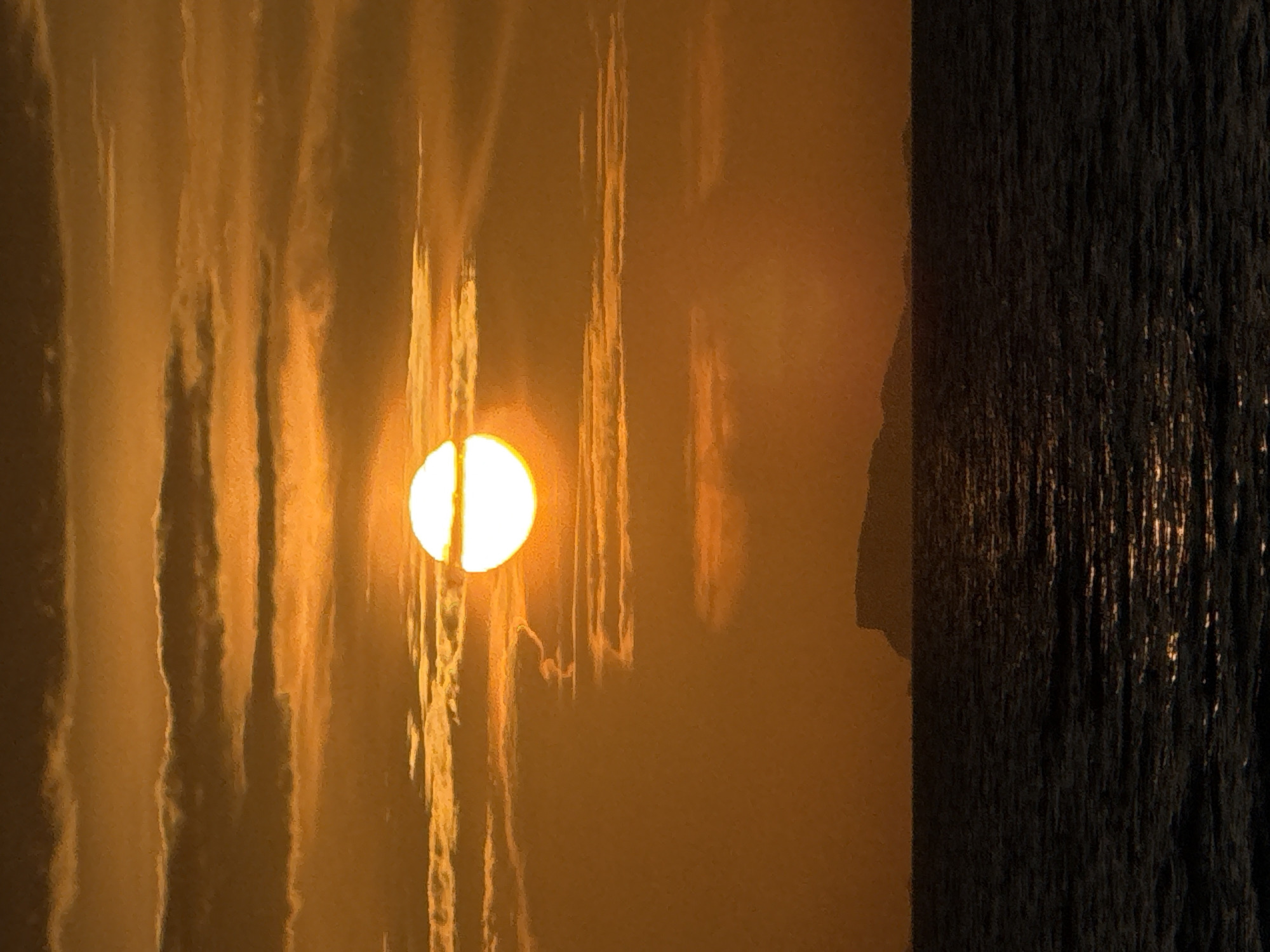 An outdoor scene shows the sun obscured by a thin line of clouds. The sky is a uniform orange, and on the right of the scene is a dark, vertical object with a textured surface. There is no indication of people, actions, or interactions in the image. The location is Baan Tai, Thailand.