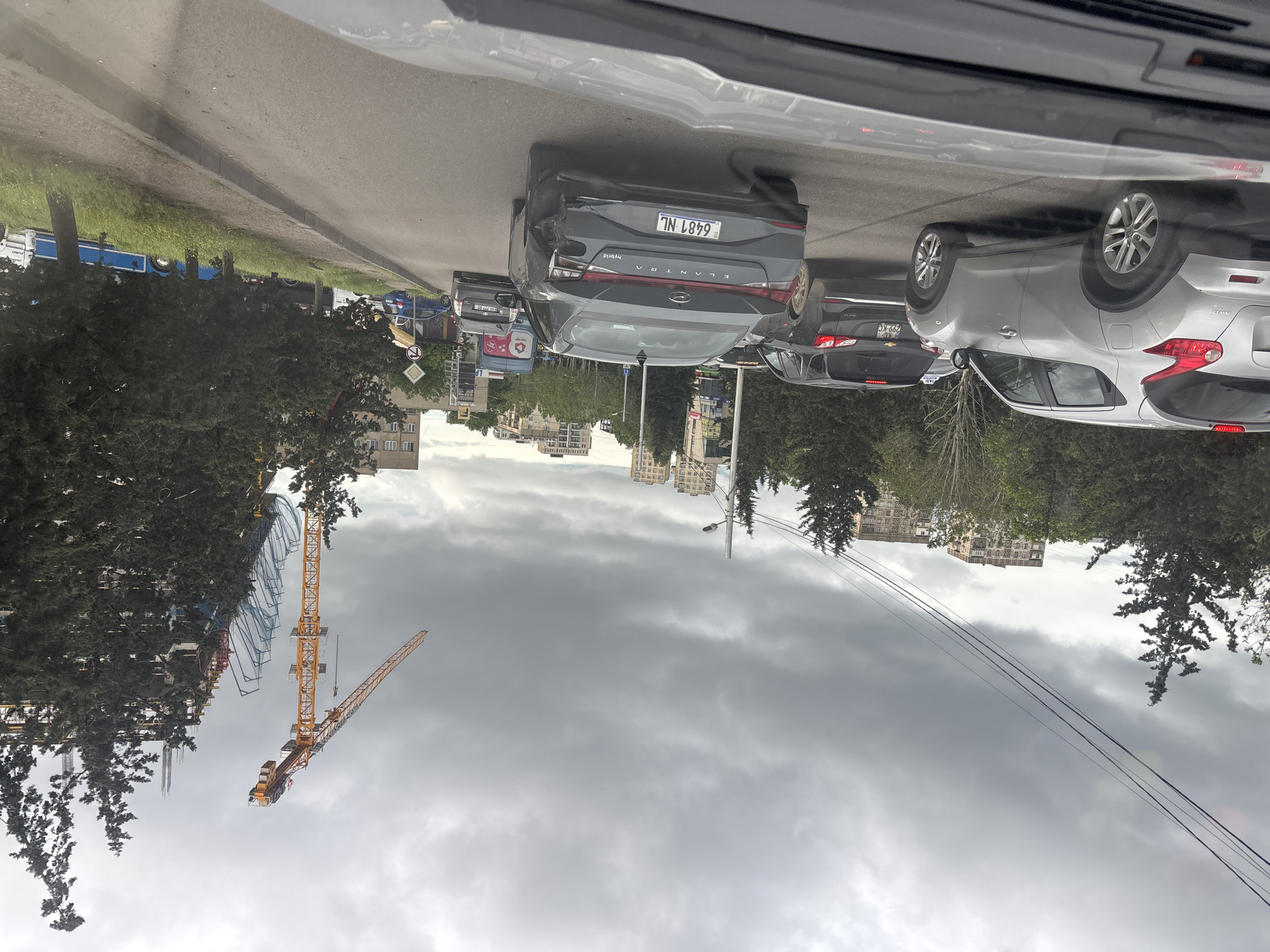 The image depicts a traffic jam on a city street, likely in Tbilisi, Georgia, under a cloudy sky. There are several vehicles visible, including a dark gray Hyundai Elantra Hybrid with license plate number 6481 NL, and a silver SUV. A construction site with a tall yellow crane is visible on the left side of the street. Buildings and power lines are present in the background. The view is from inside a vehicle, through the windshield.
