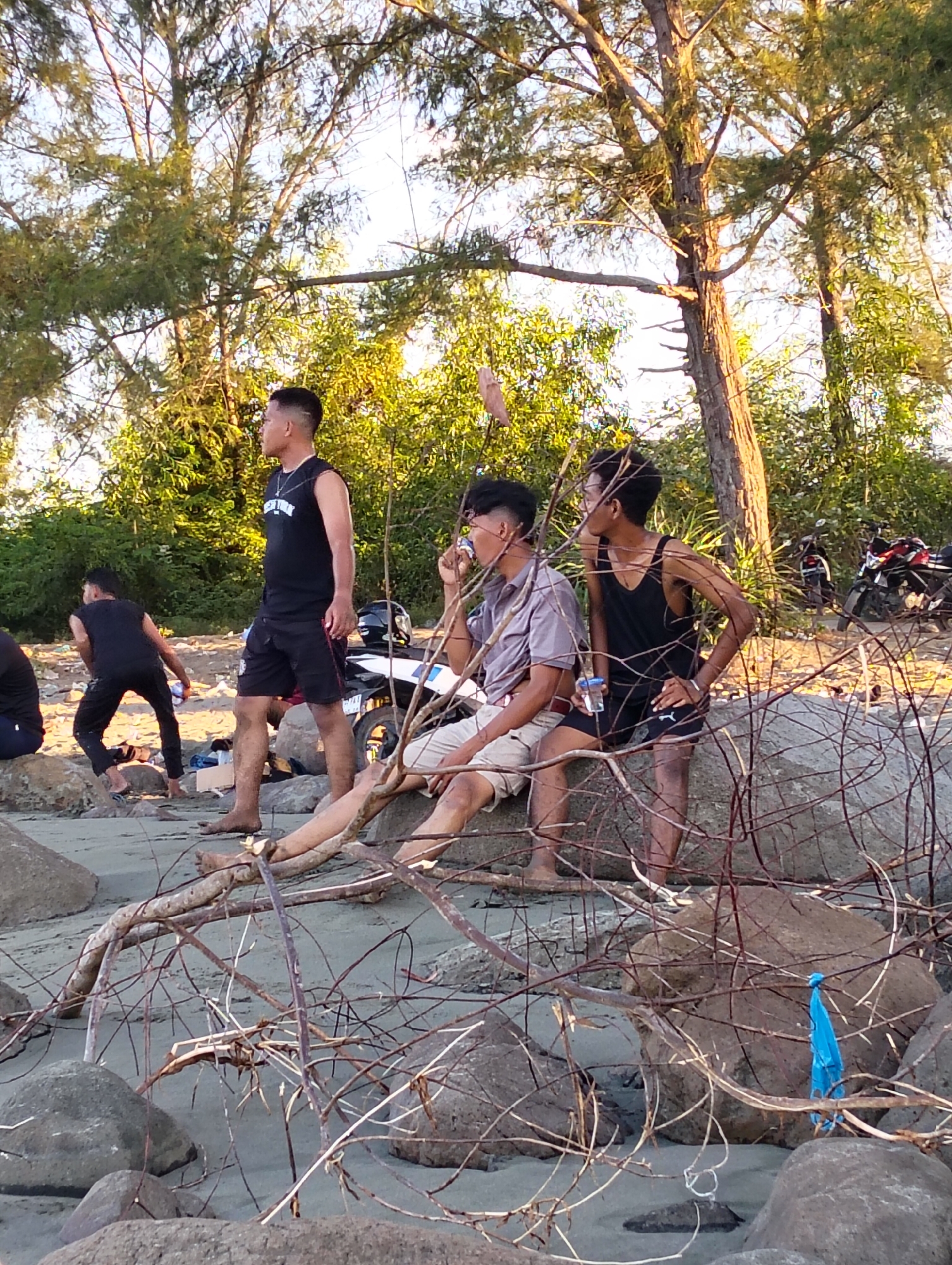The image captures a scene on a sandy beach with scattered rocks and driftwood, likely near Benteng, Indonesia. The sun is setting, casting a warm glow on the trees and vegetation in the background. In the foreground, several young men are present.
One man, wearing a black sleeveless shirt and black shorts, stands looking to his left. He has a necklace on and his hair is short and styled. To his right, another man, wearing a black tank top and black shorts, is seated on a rock, holding a clear cup. He also has short, styled hair and is wearing glasses. Further to the left, a man in a black sleeveless shirt and black shorts is captured in mid-action, appearing to be throwing something or playing a game on the sand. He is crouched down. Another individual, whose body is partially visible, is seated on a rock to the far right, wearing light-colored shorts.
In the mid-ground, a white motorcycle with red accents is parked. The beach itself is sandy with some debris and scattered rocks. The environment is natural, with trees and foliage forming a backdrop to the human activity. The overall impression is a casual gathering of friends by the sea during the late afternoon.
