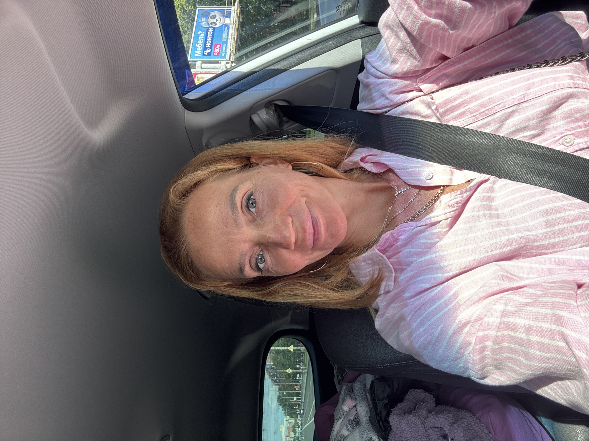 A woman with blonde hair and blue eyes smiles at the camera, taking a selfie inside a car. She is wearing a pink and white striped button-down shirt and several necklaces, including a cross pendant. A black seatbelt is fastened across her body. In the background, the car's interior is visible, along with a billboard outside the window that reads "Мебель? Нонтон" (Furniture? Nonton) and shows a cartoon panda. The view through the rear window shows a city street with buildings and trees. The scene appears to be in Saint Petersburg, Russia, given the presence of a billboard with Russian text and the architectural style of the buildings.