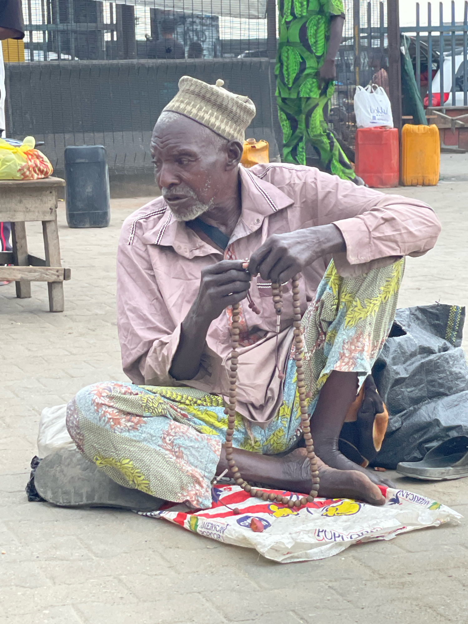 In Ajah, Nigeria, a man sits cross-legged on the ground, holding a string of wooden prayer beads. He wears a light pink button-down shirt and a patterned fabric wrapped around his lower body. His head is covered with a textured, beige cap. The ground is paved with concrete blocks, and a worn rug is spread beneath him. To his left, a wooden stool supports a yellow plastic bag with items inside. A black plastic barrel stands behind the stool. In the background, a person in a vibrant green garment with a black and green leaf pattern walks away from the camera. Further back, various containers, including red and yellow plastic drums, and a white shopping bag are visible. A dark tarp and a pair of black sandals are situated to the man's right. The scene appears to be an outdoor urban setting, possibly a market or street side.