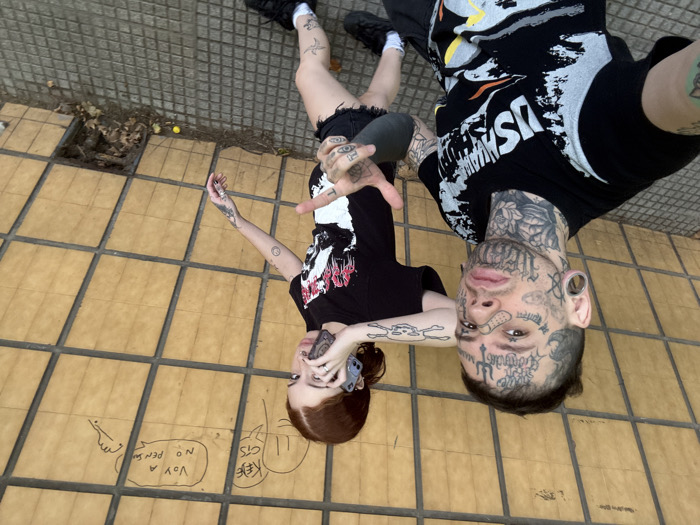 The image captures an overhead view of two individuals lying on a tiled outdoor surface, looking up at the camera. The perspective makes them appear as if they are lying on their backs with their heads pointing towards the viewer.
The person in the foreground, on the right side of the image, is a male with extensive tattoos covering his face, neck, and arms. He has a dark, short hairstyle and large ear gauges. There is a small band-aid visible below his right eye. He is wearing a black t-shirt with a white and gray graphic design that includes the partial word "US ARMY" and other abstract shapes. His left arm, also heavily tattooed, is visible, and his hand is making a gesture with his index finger pointing upwards and thumb extended.
The second person, positioned behind and slightly to the left of the first, is a female with long, reddish-brown hair. She is also heavily tattooed on her arms and legs. She is wearing a black t-shirt with a red and white graphic design, and black denim shorts. Her legs, crossed at the ankles, are visible in the upper left corner of the frame. Her right hand, also tattooed, is holding a black smartphone up towards the camera.
The ground beneath them is covered with light brown square tiles separated by dark gray grout lines. To the left and below the female's head, there are several markings drawn directly onto the tiles. One speech bubble contains the partially legible text "VOY A NO PENSA" (roughly translating to "I'm going to not think") next to a drawn hand. Another bubble has what appears to be "KEE C STS" and a third contains "LL".
In the upper left corner, beyond the paved area, there is a patch of uneven ground with dried leaves and some debris. A small, round, yellow object is visible on the ground in this area. The overall aesthetic of the individuals, with their numerous tattoos and dark clothing, suggests an alternative or edgy style. The scene appears to be taking place outdoors during daylight hours.
