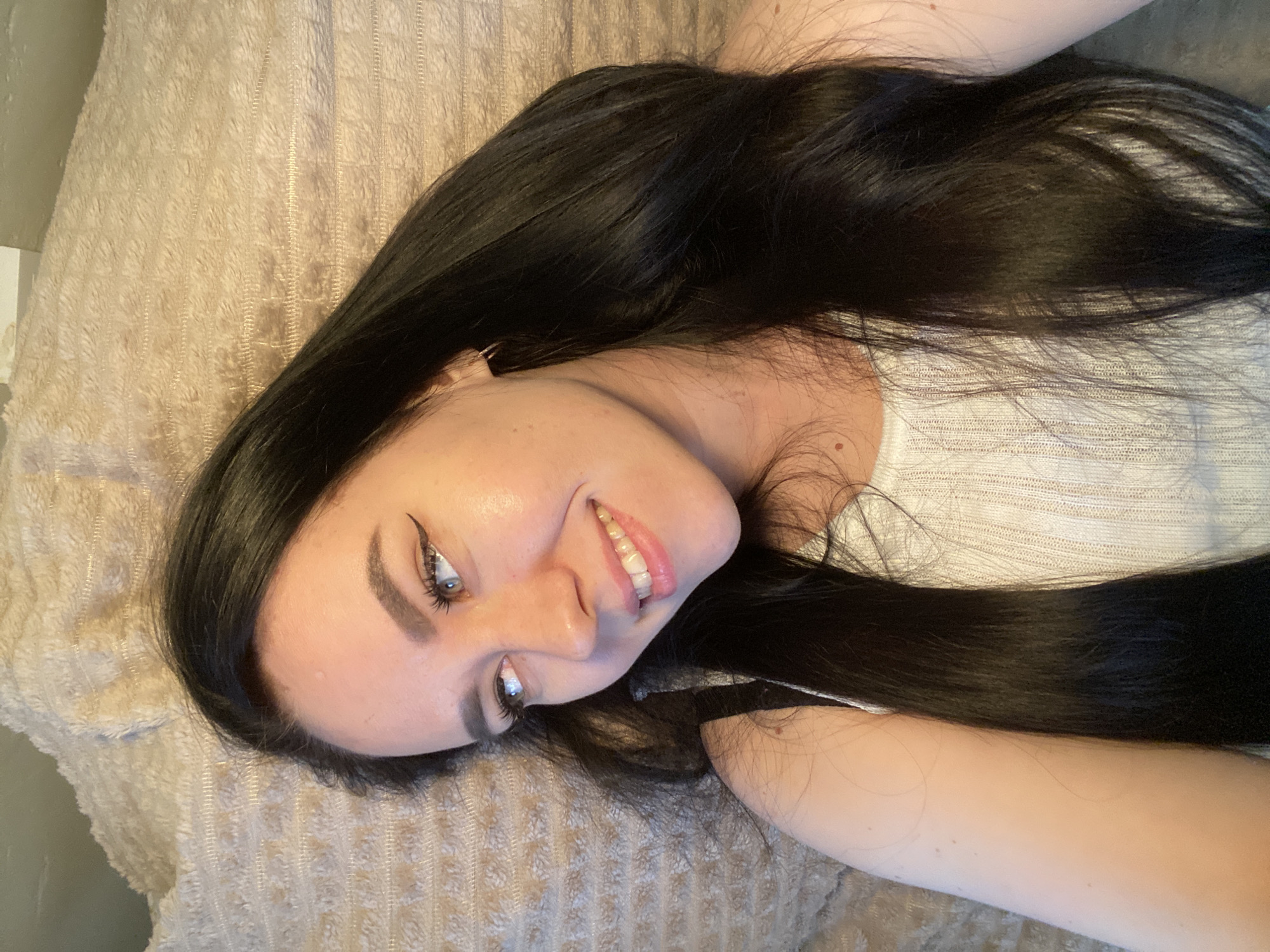A woman with long, dark hair is lying on a beige textured surface. She is wearing a white top and has eyeliner on. She is smiling and looking up at the camera. The location is not discernible from the image.