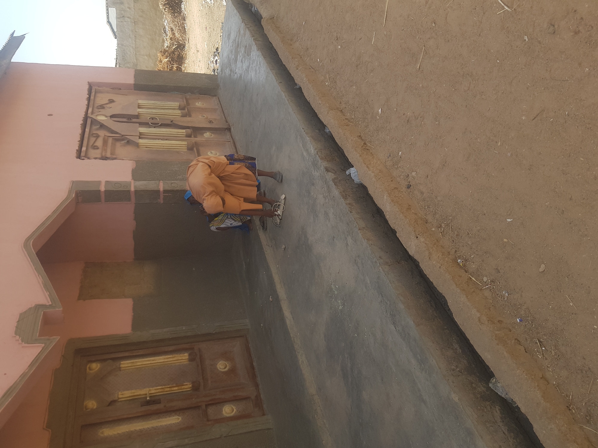 The image shows the exterior of a building in Dutsinma B, Nigeria. The building is painted peach with brown accents. There are two doors visible, one at ground level and another above it. A person wearing a tan dress or robe is bent over, seemingly interacting with something on the concrete walkway in front of the building. A ditch runs parallel to the walkway, separating it from a dirt area. In the background, there is what appears to be construction material or debris. The scene is well-lit by daylight.