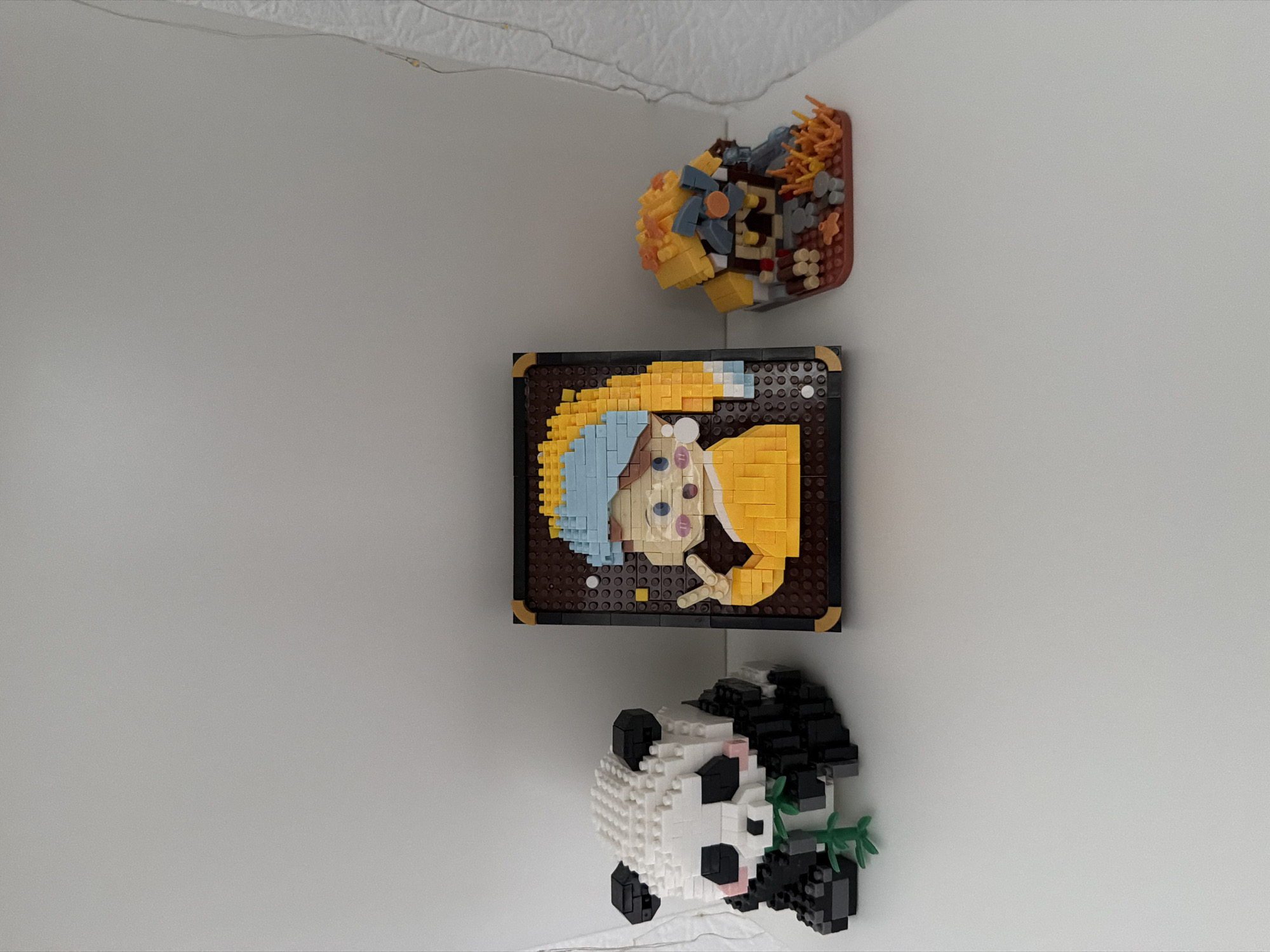The image shows three small figurines made of building blocks, likely from a toy set, displayed on a white shelf against a plain white background. On the left is a panda figurine eating bamboo. In the center is a framed piece depicting a stylized portrait of a young girl in a yellow outfit with a blue head covering, giving a peace sign. On the right is a small, rustic building with a thatched roof, possibly a cabin or a shack, situated on a base with rocks and foliage. The overall setting appears to be an indoor display, and there is no indication of the city of Kyiv, Ukraine, or any specific urban environment.