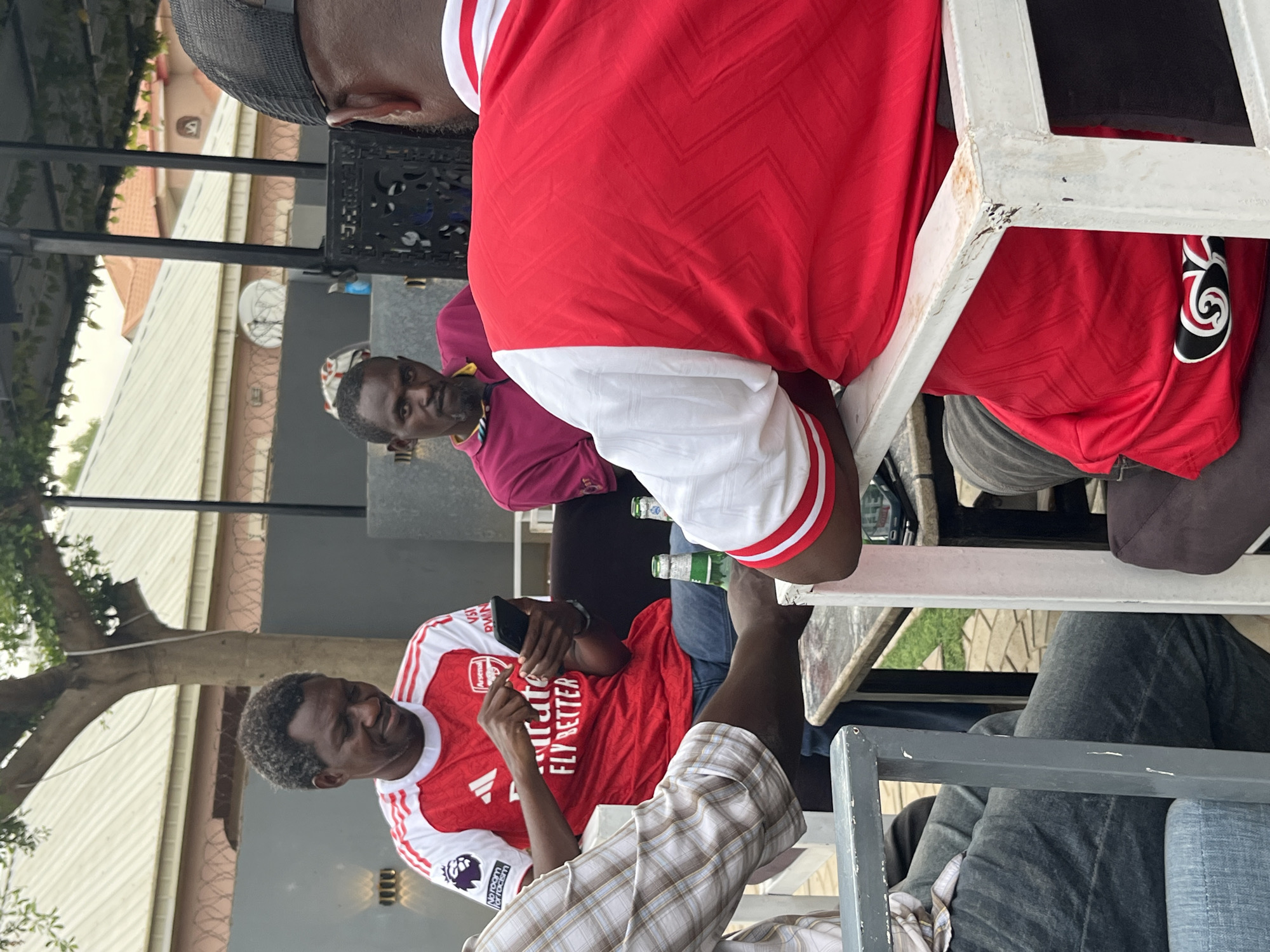 In Gura Top, Nigeria, a group of men are gathered outdoors. Several are wearing red and white soccer jerseys, one of which has "Emirates Fly Better" printed on it. One man is looking at a phone while raising a finger. A bottle of beer is visible on the table. The location appears to be a restaurant or gathering place with outdoor seating.