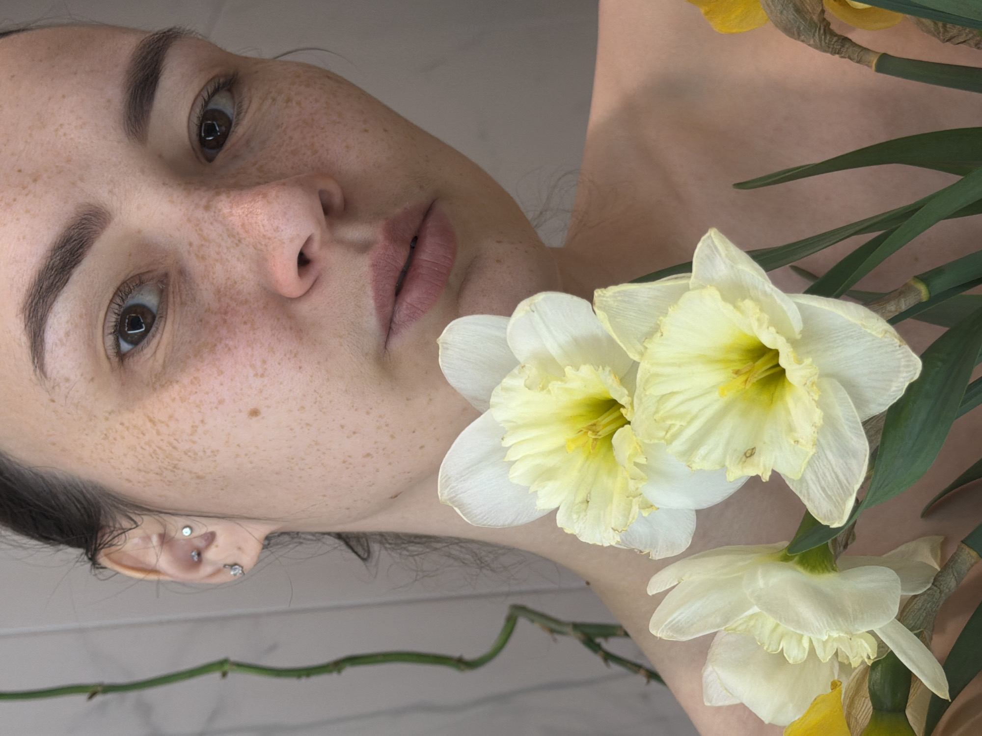 A young woman's face is the primary focus of the image, surrounded by white and yellow daffodils. She has light skin with freckles, dark eyebrows, and brown eyes. She has multiple piercings in her ear. The background is a light, neutral color, possibly a wall. The daffodils are in bloom and appear fresh. The location in the city of Chaikovsky, Russia, is not discernible from the visual information provided in the image.
